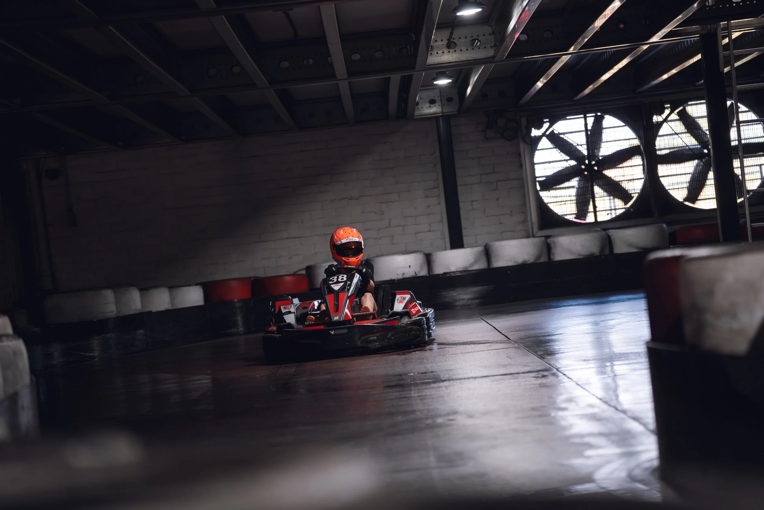 A person wearing a red helmet and racing suit driving a go-kart on an indoor track with safety barriers and large industrial fans on the wall.