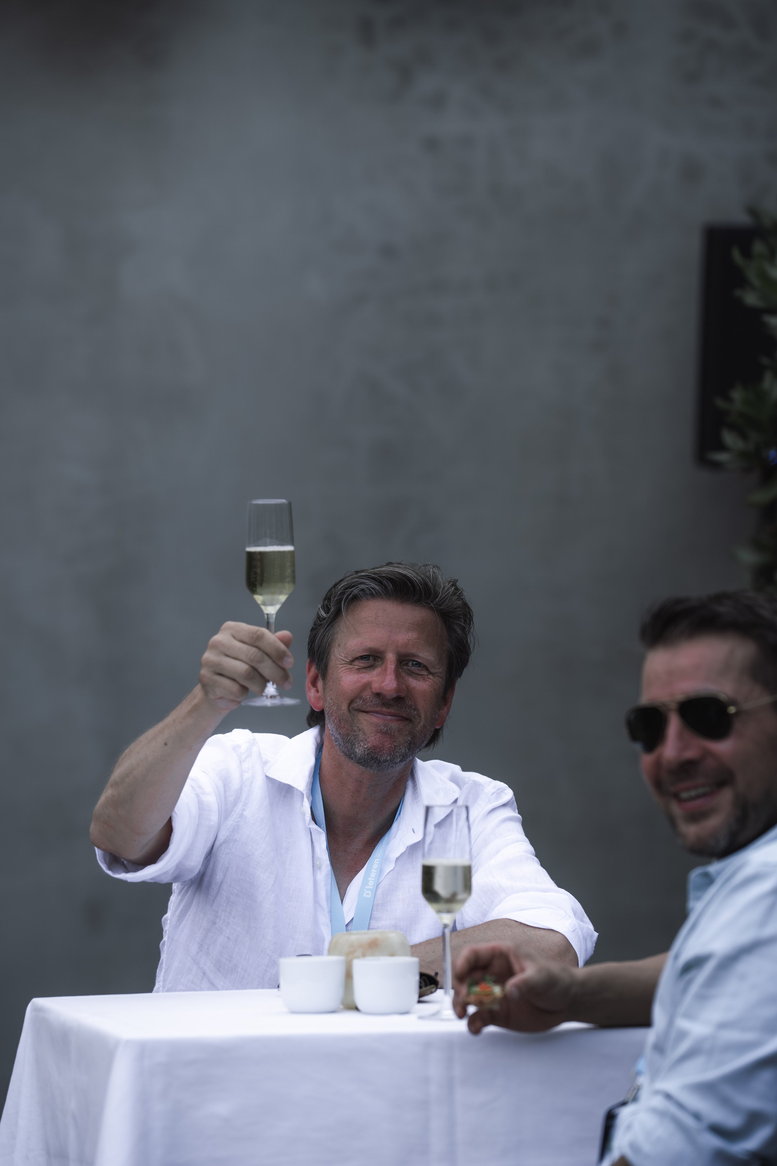 Two men sitting at a white table, smiling, holding glasses of champagne in a toast, with a dark wall in the background.