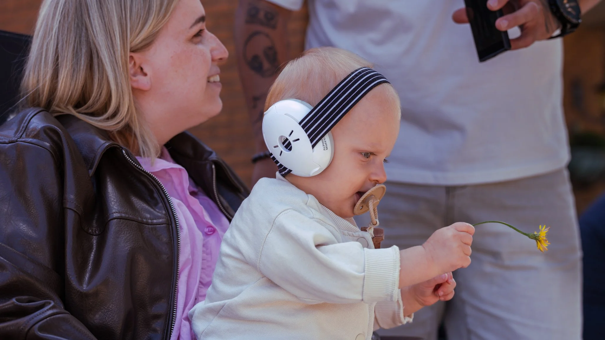 A woman with blonde hair wearing a brown leather jacket holds a toddler with a pacifier and large white headphones, while the toddler holds a yellow flower.