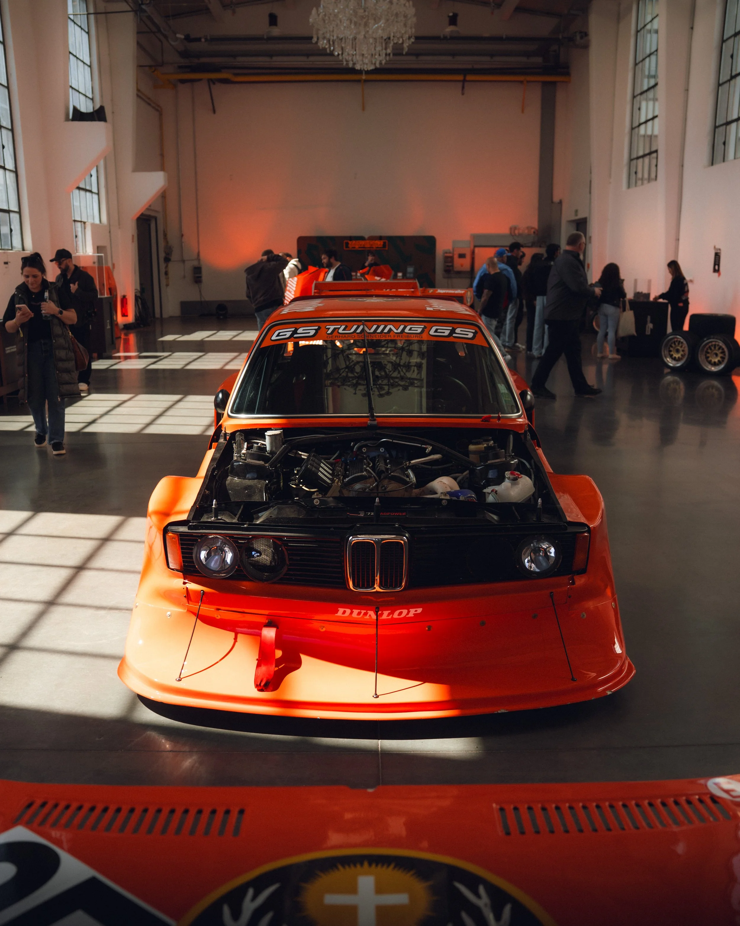 An orange vintage racing car on display at an indoor exhibition with people looking at it and a chandelier hanging from the ceiling.