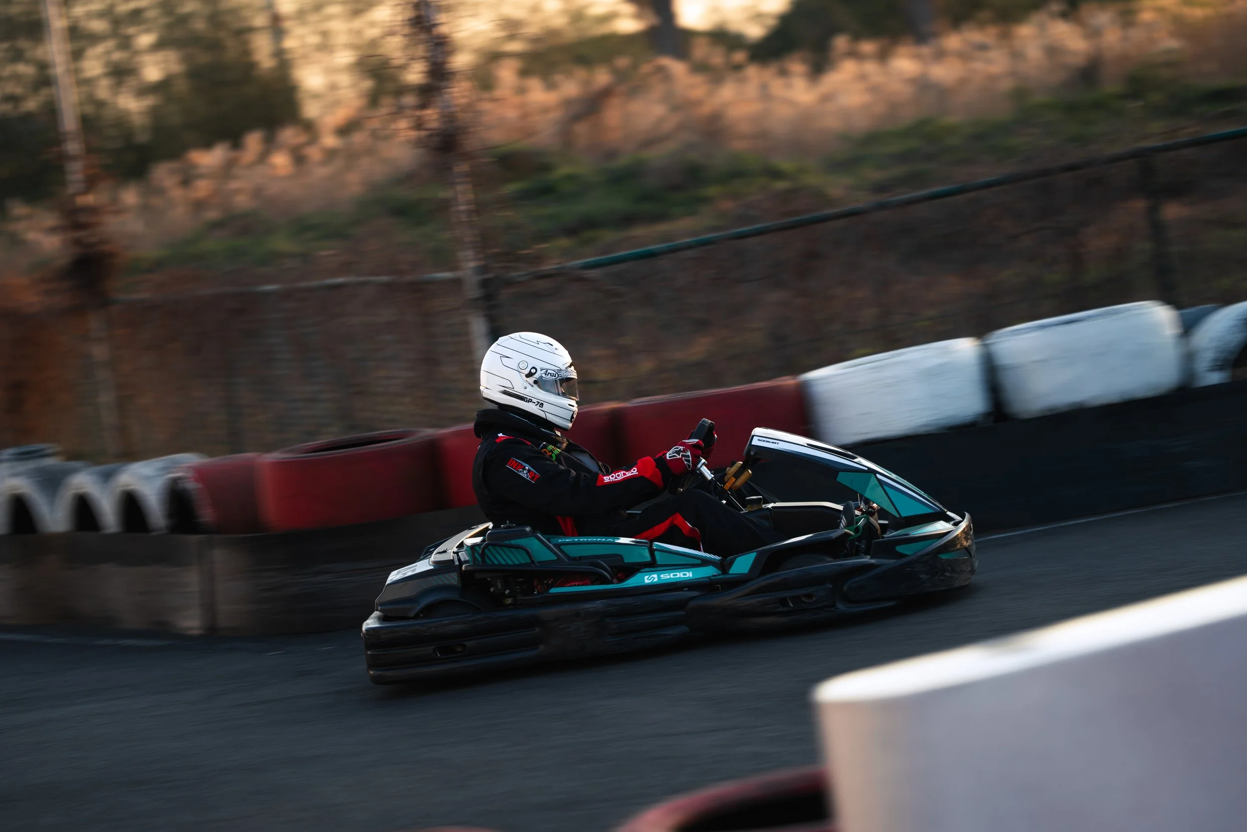 A go-kart driver in a racing suit and helmet speeding on a track, with tire barriers along the edge and a blurred background.
