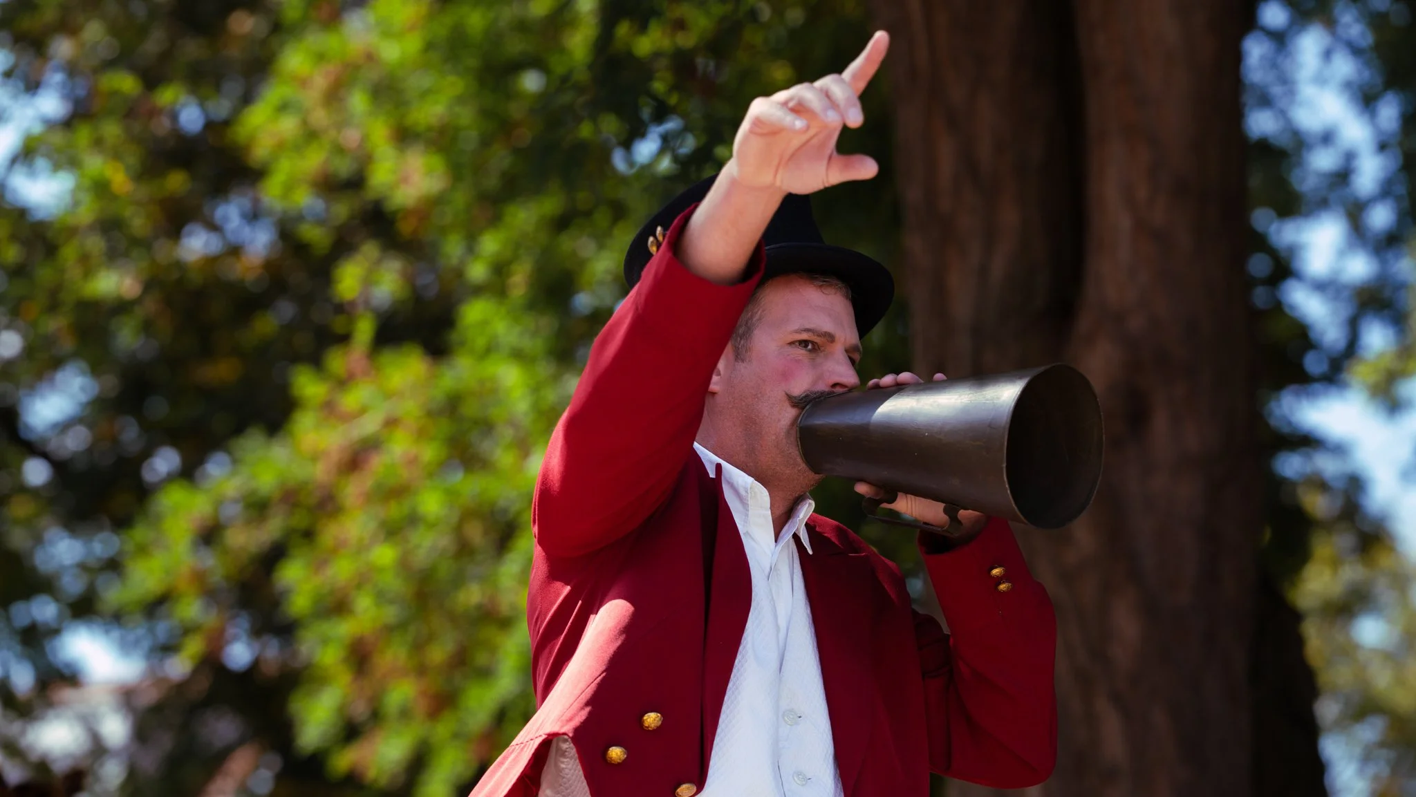 A man with a mustache and dressed in vintage clothing is speaking into a megaphone outdoors. He is pointing with his right hand against a background of green trees.
