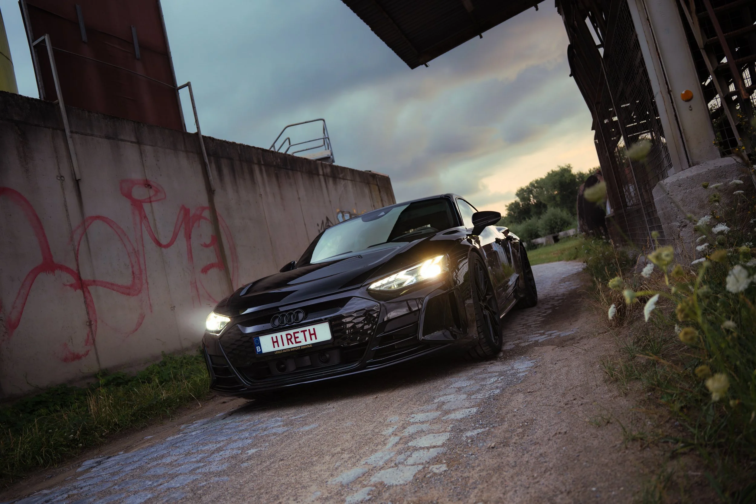 A black Audi sports car with headlights on, parked under a bridge near a graffiti-covered concrete wall, with a cloudy sky in the background.