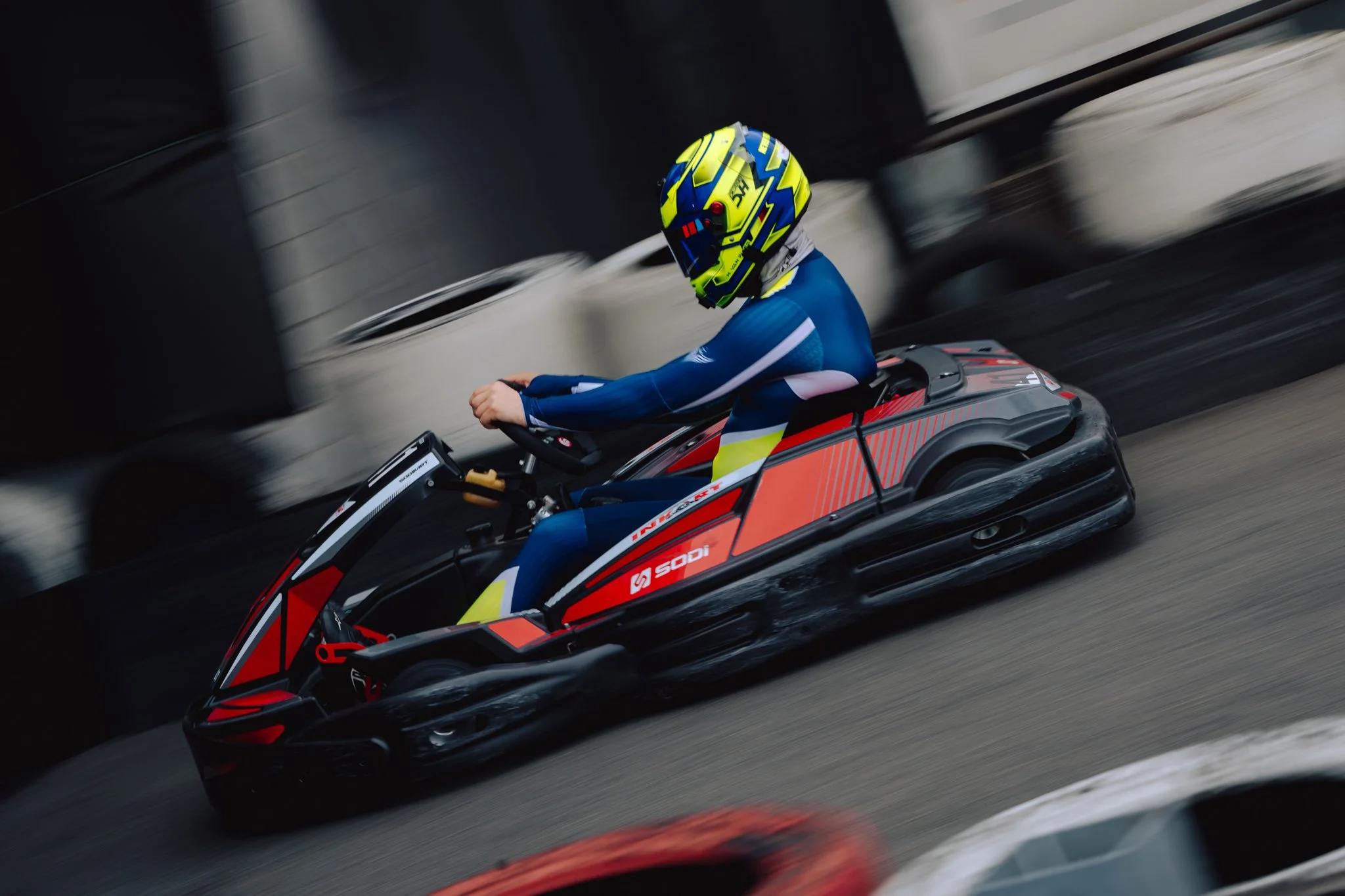 A person wearing a blue and yellow helmet and a blue racing suit is driving a black and red go-kart on a racetrack.