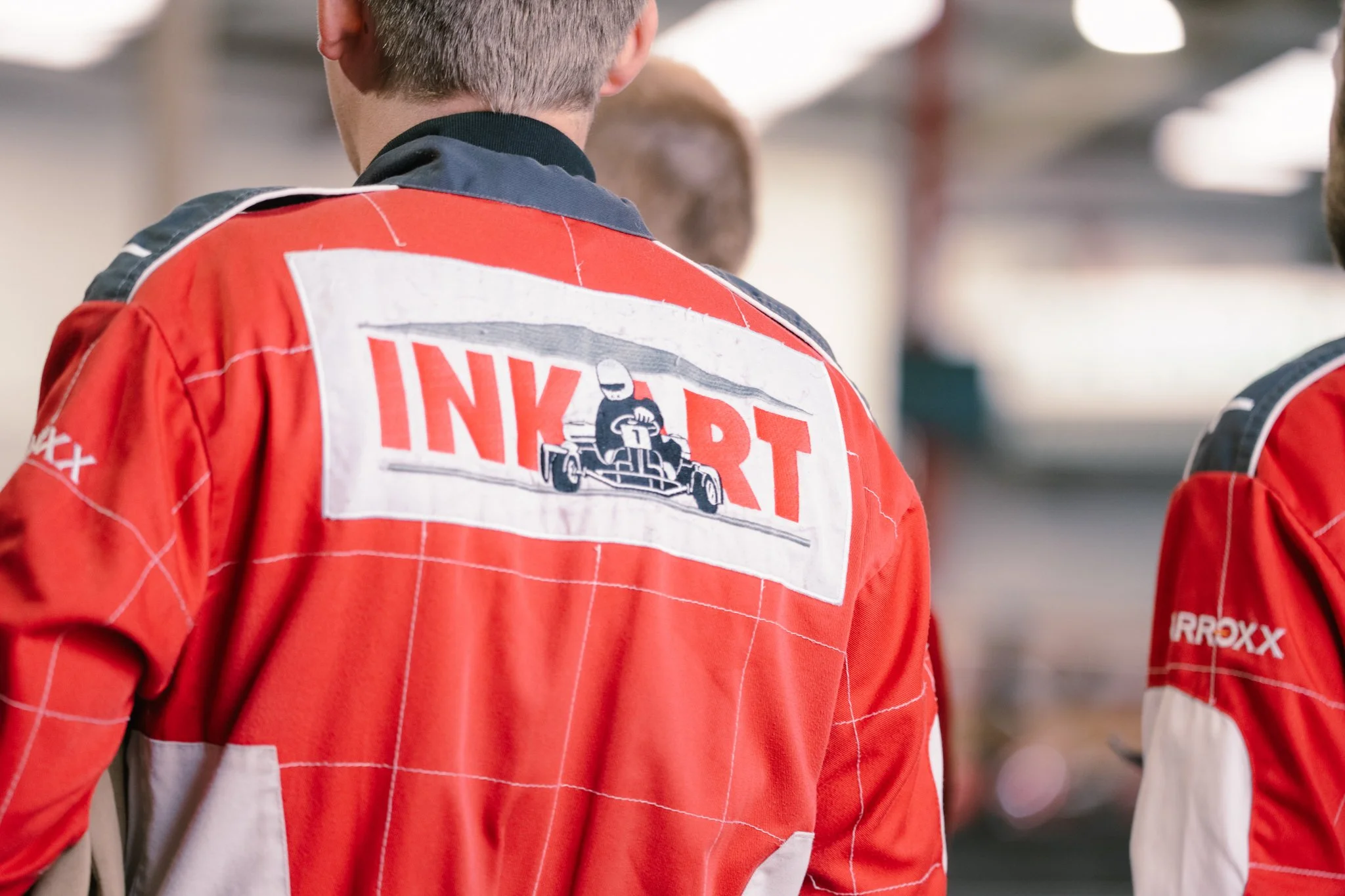 Back of a person wearing a red racing suit with a patch that says 'INKART' and depicts a go-kart driver.