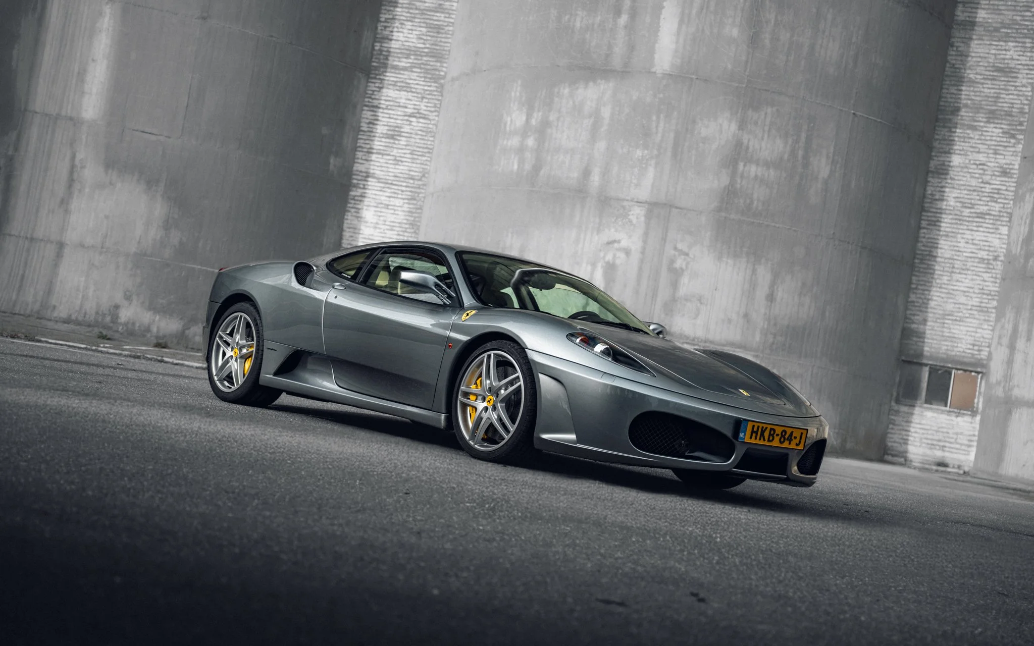 A silver Ferrari sports car parked on a paved surface next to a large concrete wall.