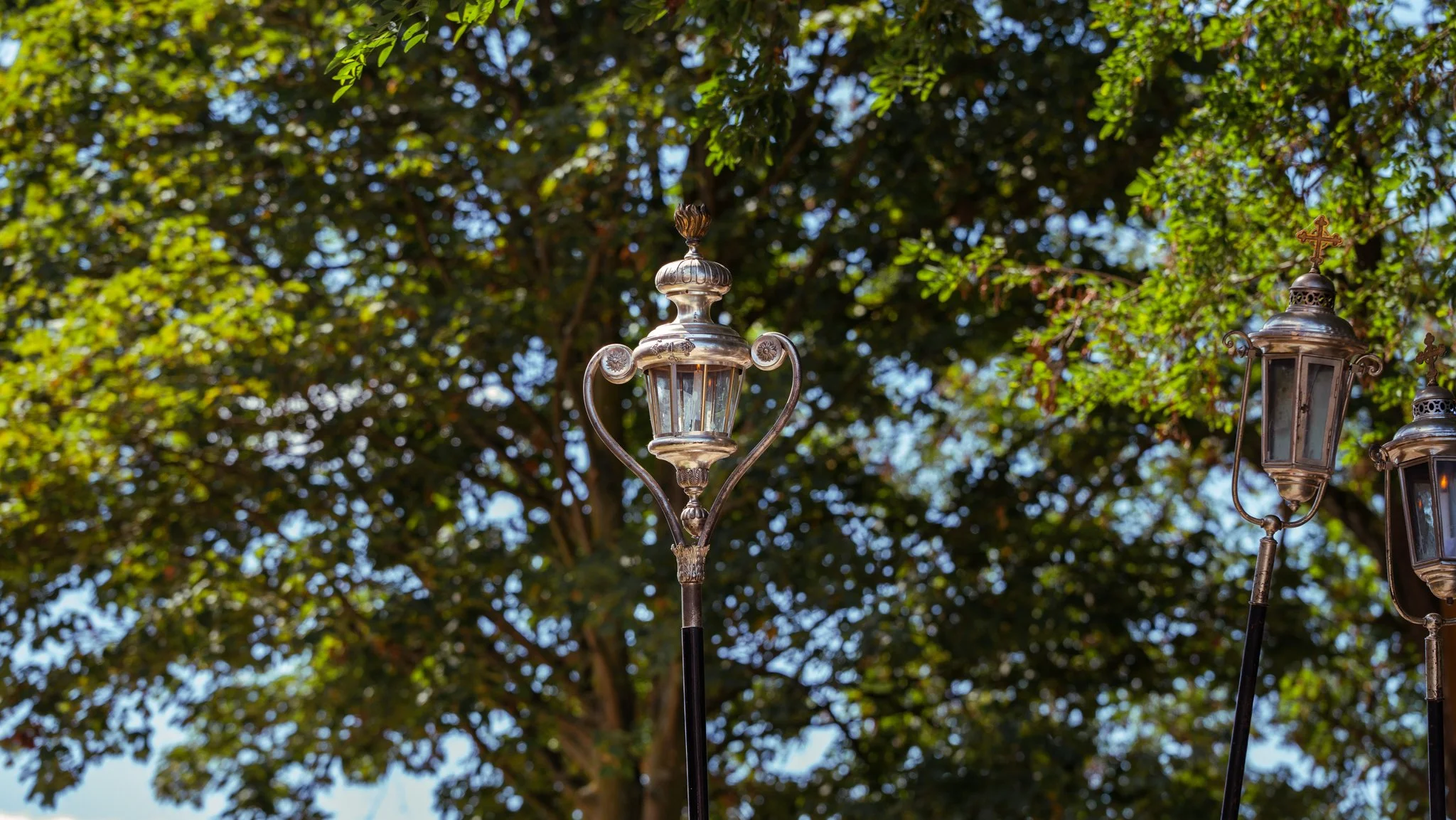 Three vintage street lamps against a background of green, leafy trees with sunlight filtering through.