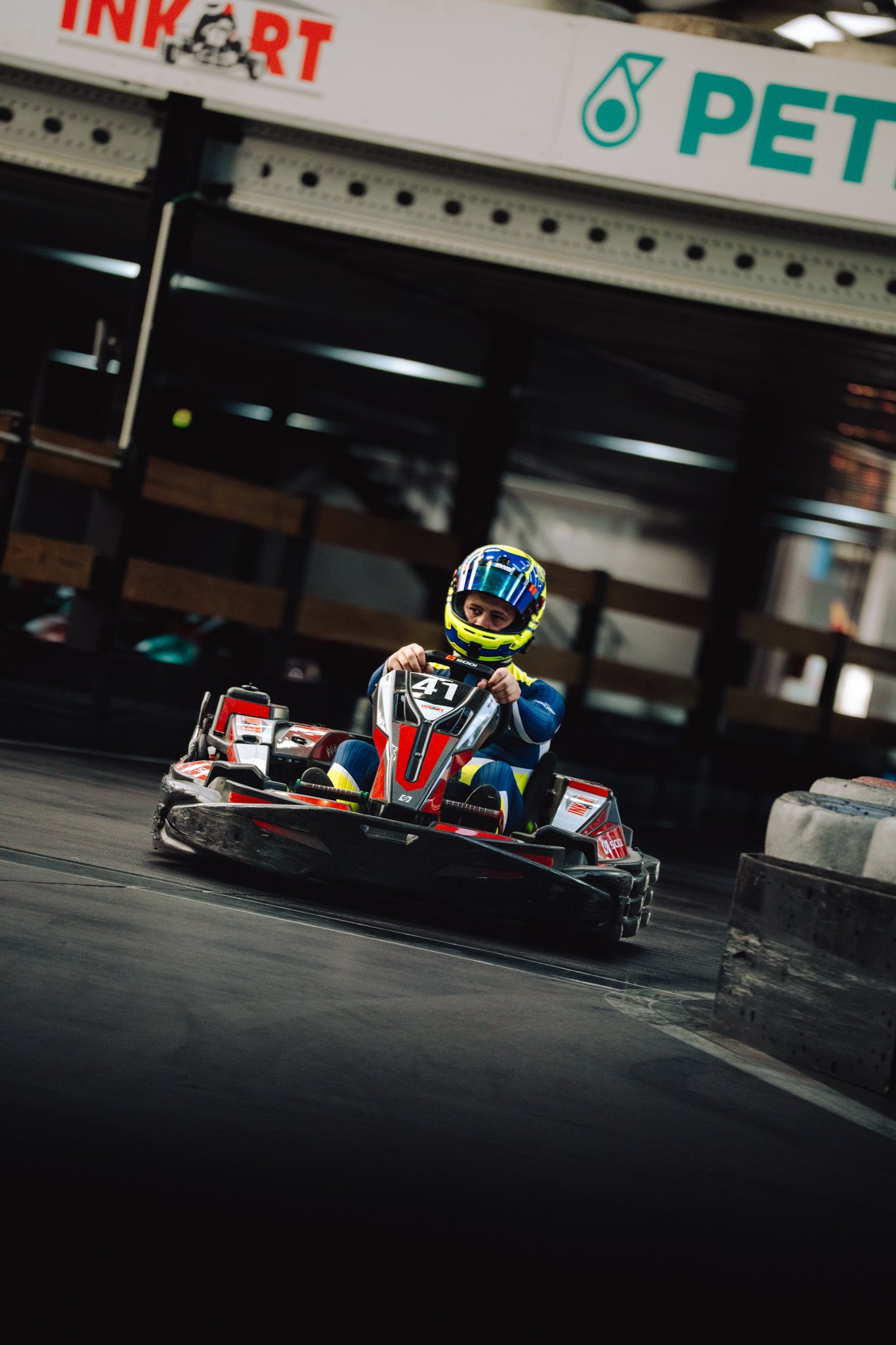 A person wearing a blue racing suit and a colorful helmet driving a go-kart on an indoor track.