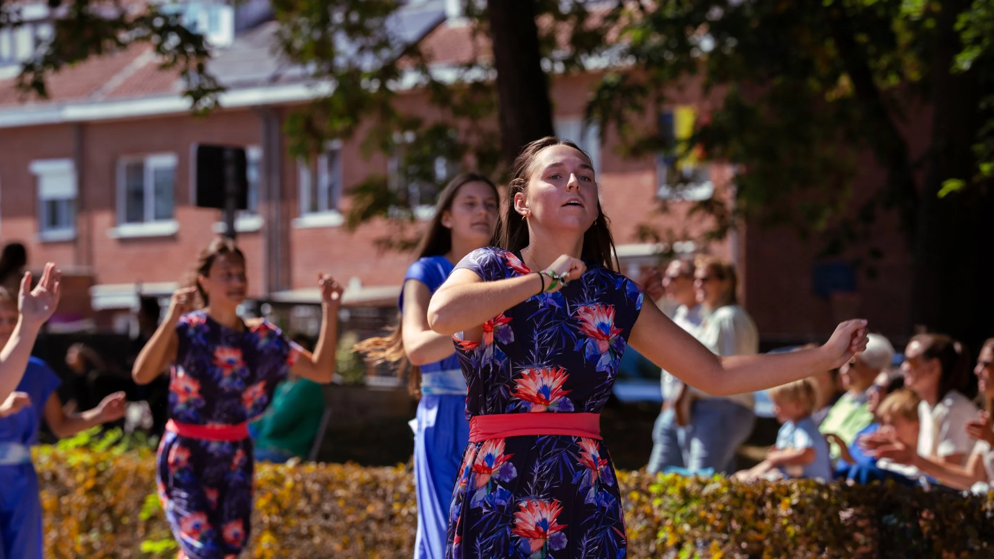 Young women dancing outdoors during a festive event with a crowd watching in the background.