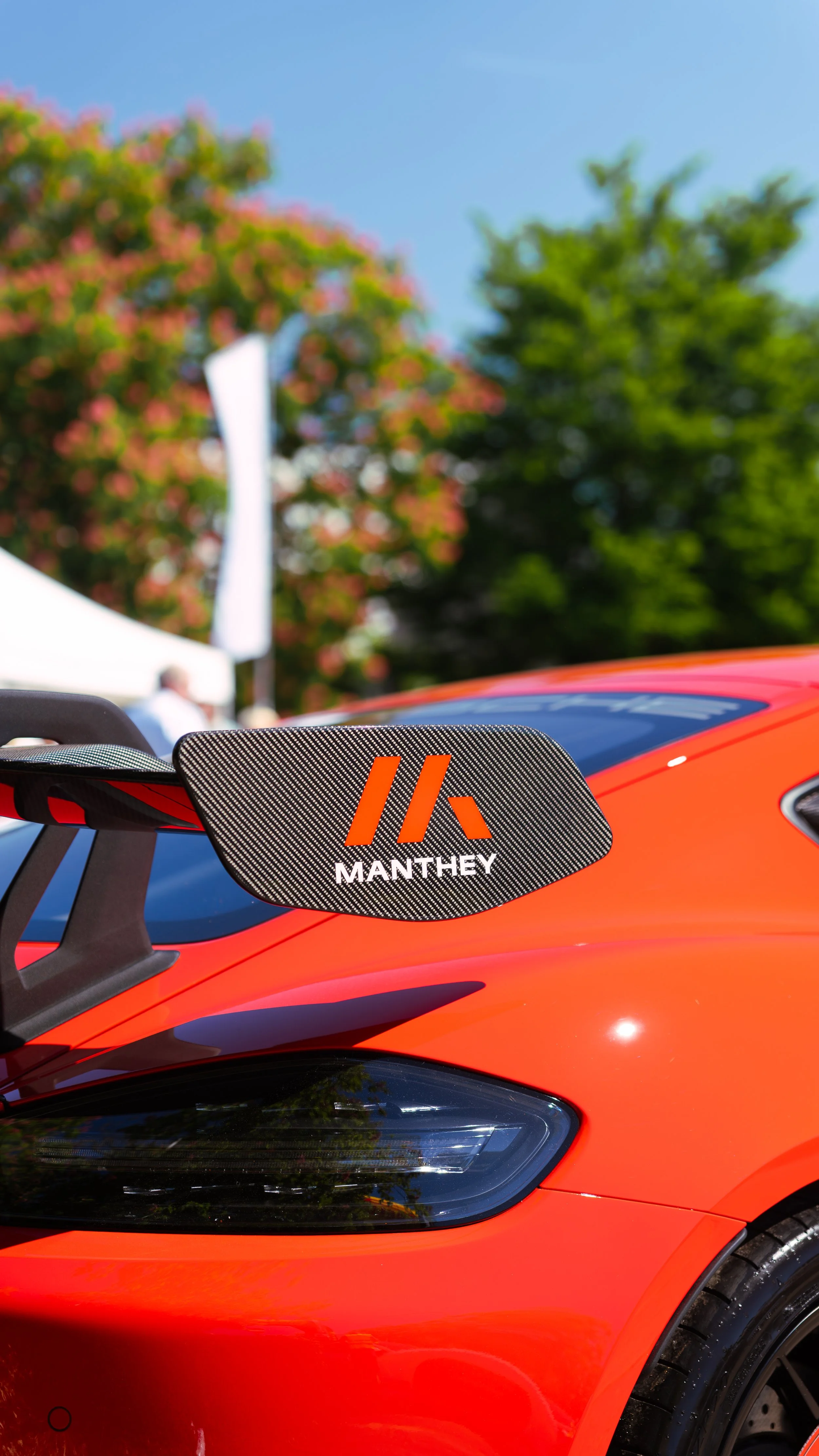 Close-up view of a red race car with a black and orange MANTHEY logo on the rear wing, parked outdoors under a blue sky with trees in the background.