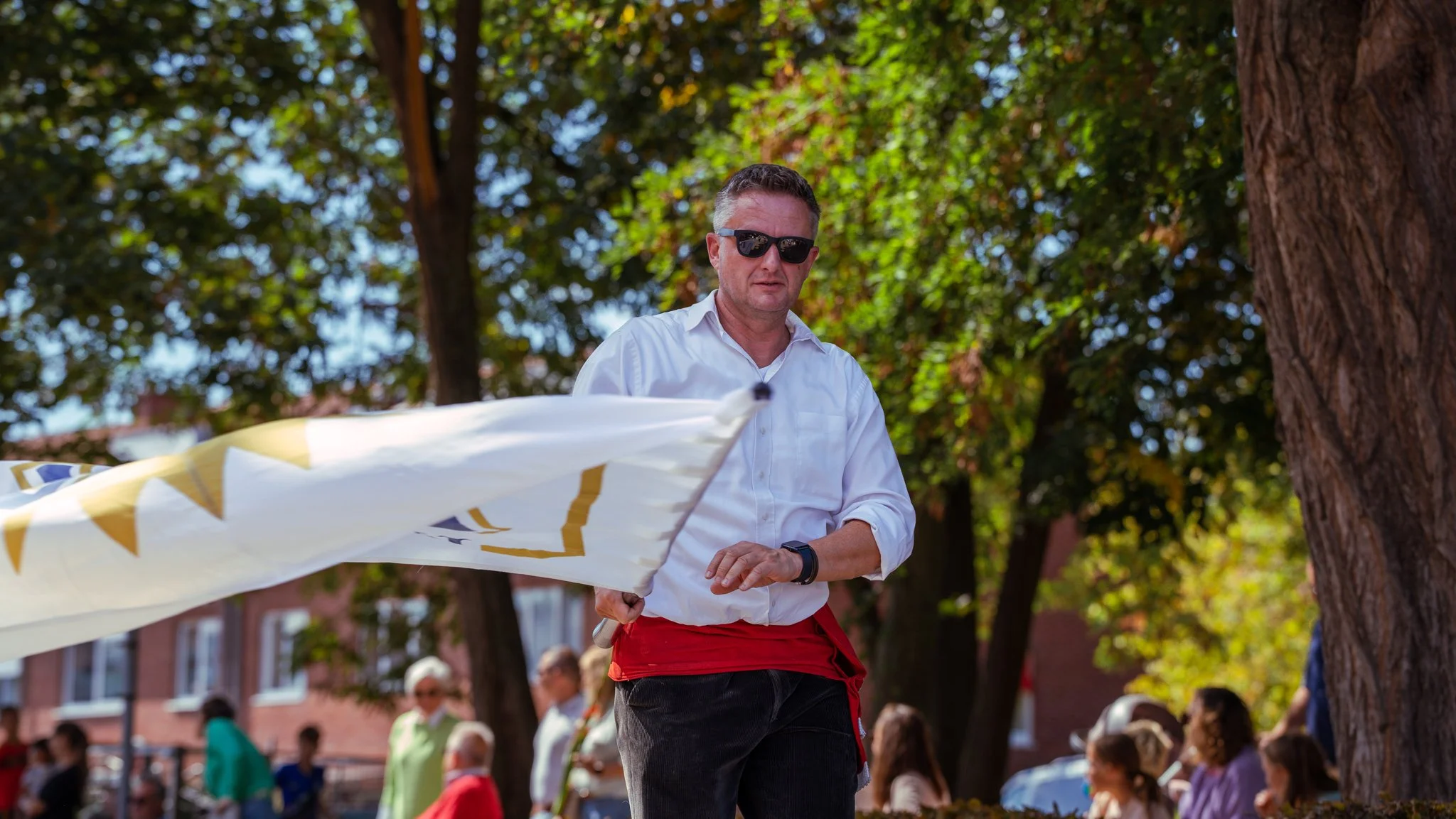 A man wearing sunglasses and a white shirt holding a flag in a park with trees and people in the background.