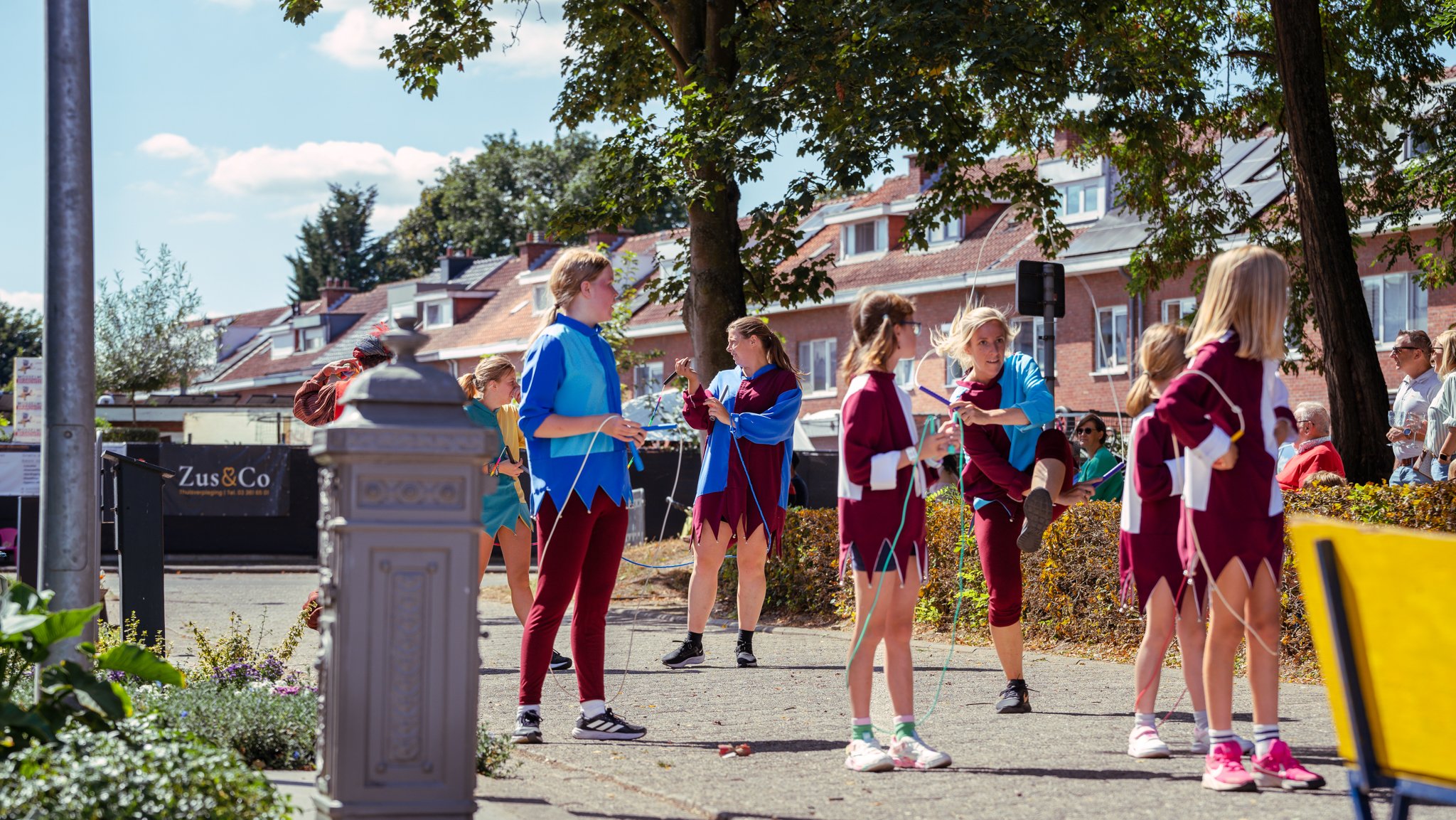 Group of children dressed as elves practicing with ropes outdoors on a sunny day, with adults sitting nearby and residential buildings in the background.
