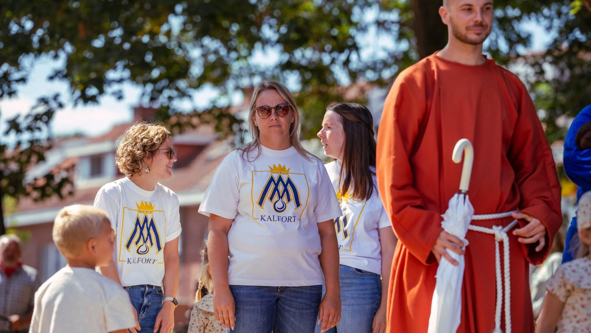 Group of people outdoors, including women wearing white t-shirts with a blue and gold logo and a woman wearing sunglasses, standing near a man in an orange robe holding an umbrella with a white handle, in front of trees and buildings on a sunny day.