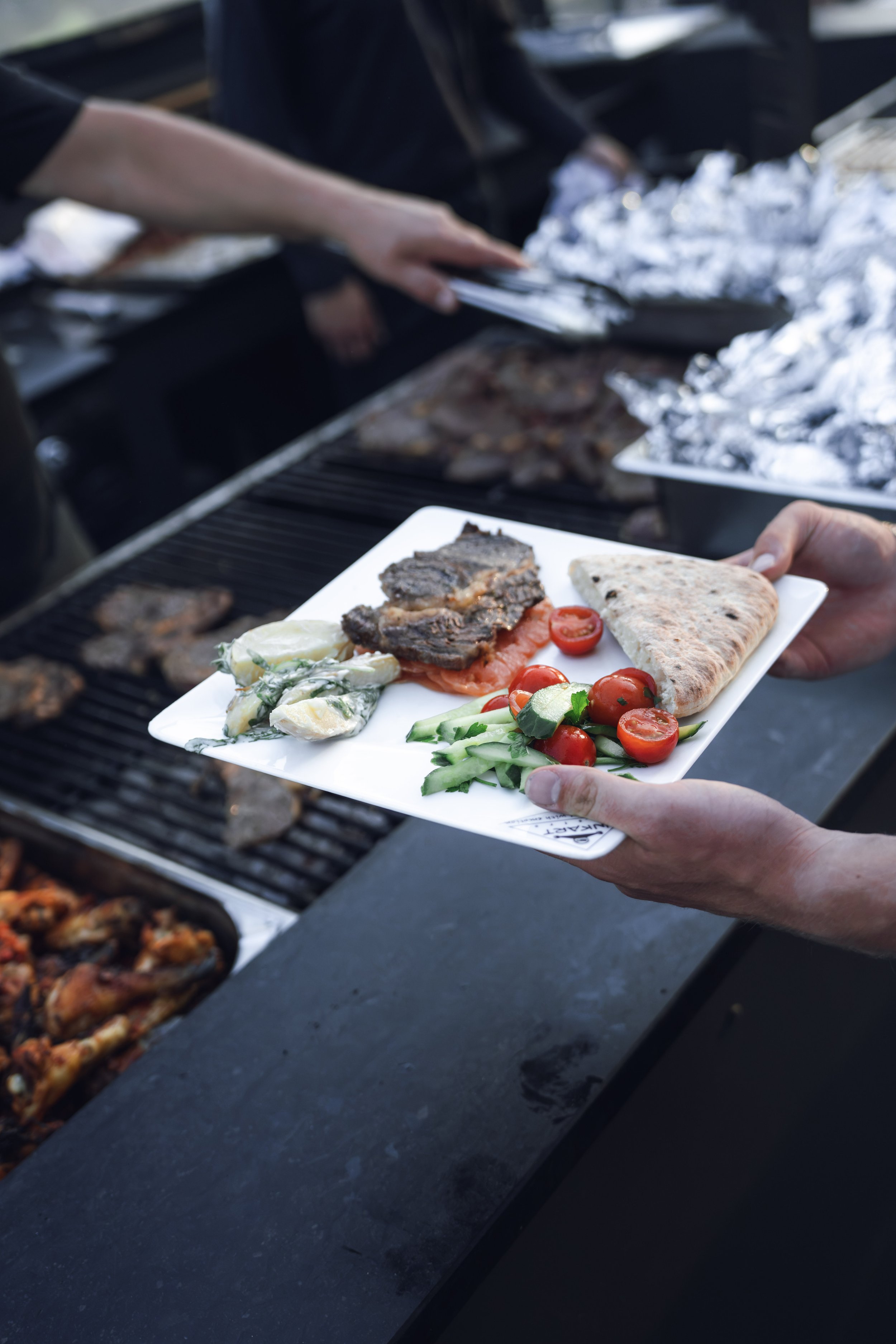 Person holds a white plate with grilled meat, cherry tomatoes, cucumber salad, potato salad, and flatbread, at a barbecue grill with foil-covered food in the background.