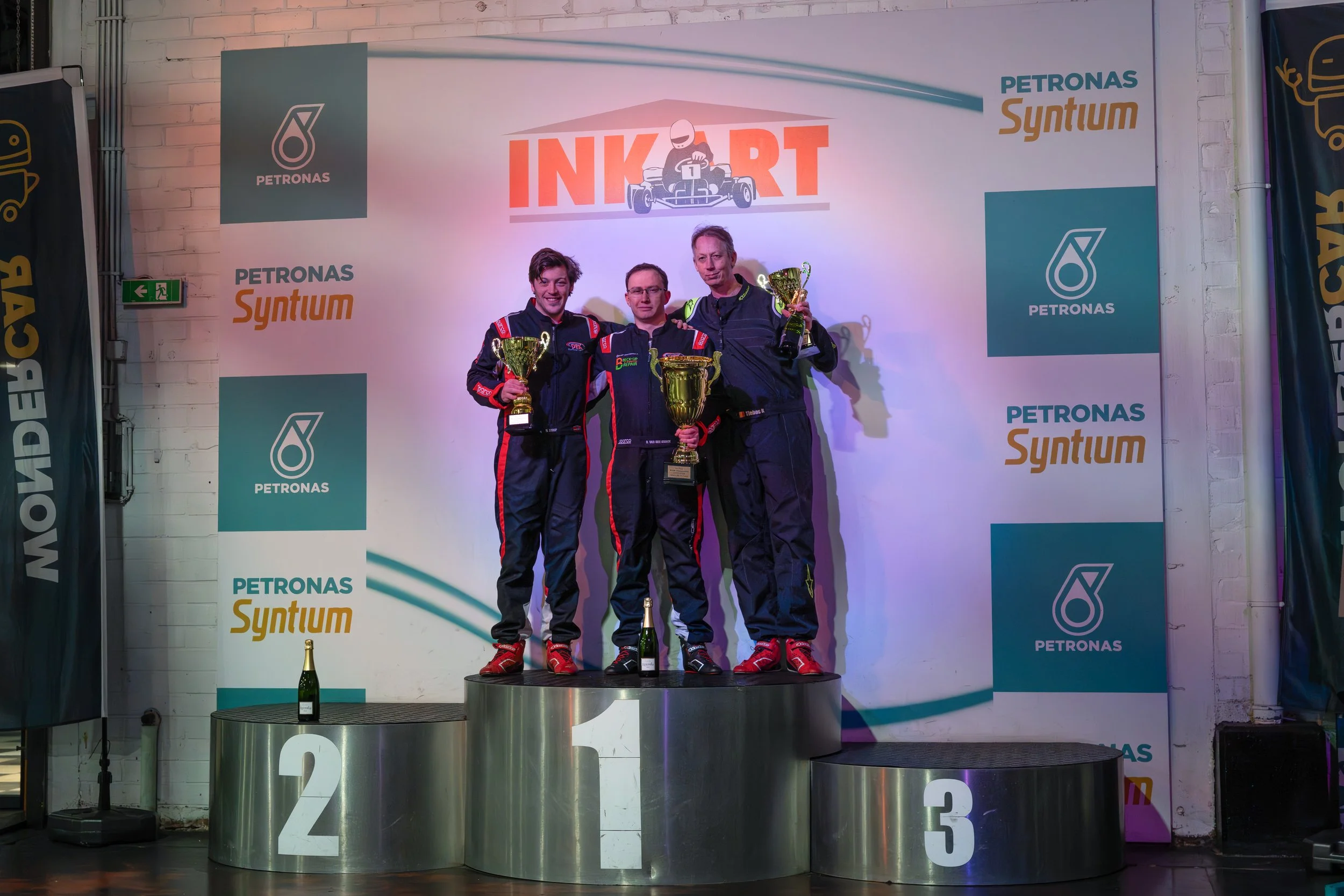 Three race car drivers standing on a Winners' Podium holding trophies, celebrating their victory. The podium is numbered 1, 2, and 3, with the first-place winner in the center holding a large cup. The background features a banner with the logo of Pet