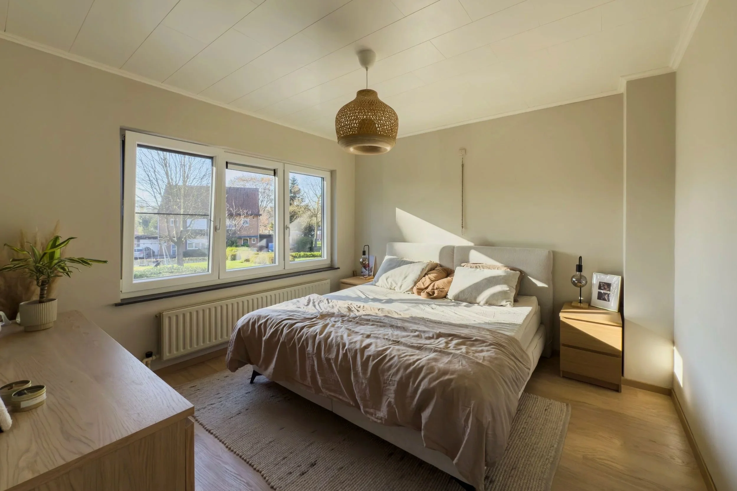 Bright bedroom with a bed, pillows, nightstands, a large window, and a hanging light fixture.