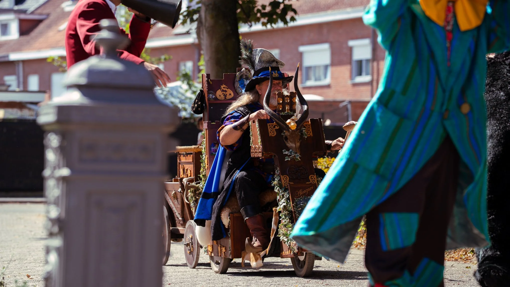 A person dressed as a medieval or fantasy character, with a horned helmet and blue cloak, seated in a decorated cart with a bull skull on it. The scene is outdoors on a sunny day, with other people in colorful costumes and a brick building in the bac
