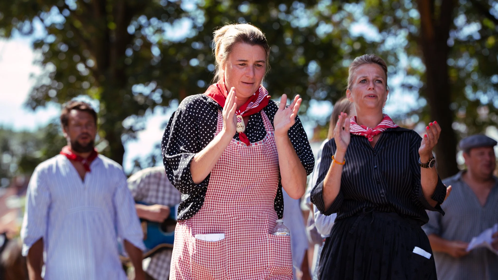 Group of people outdoors, with two women in foreground appearing emotional and crying, others in background, trees and blue sky visible.