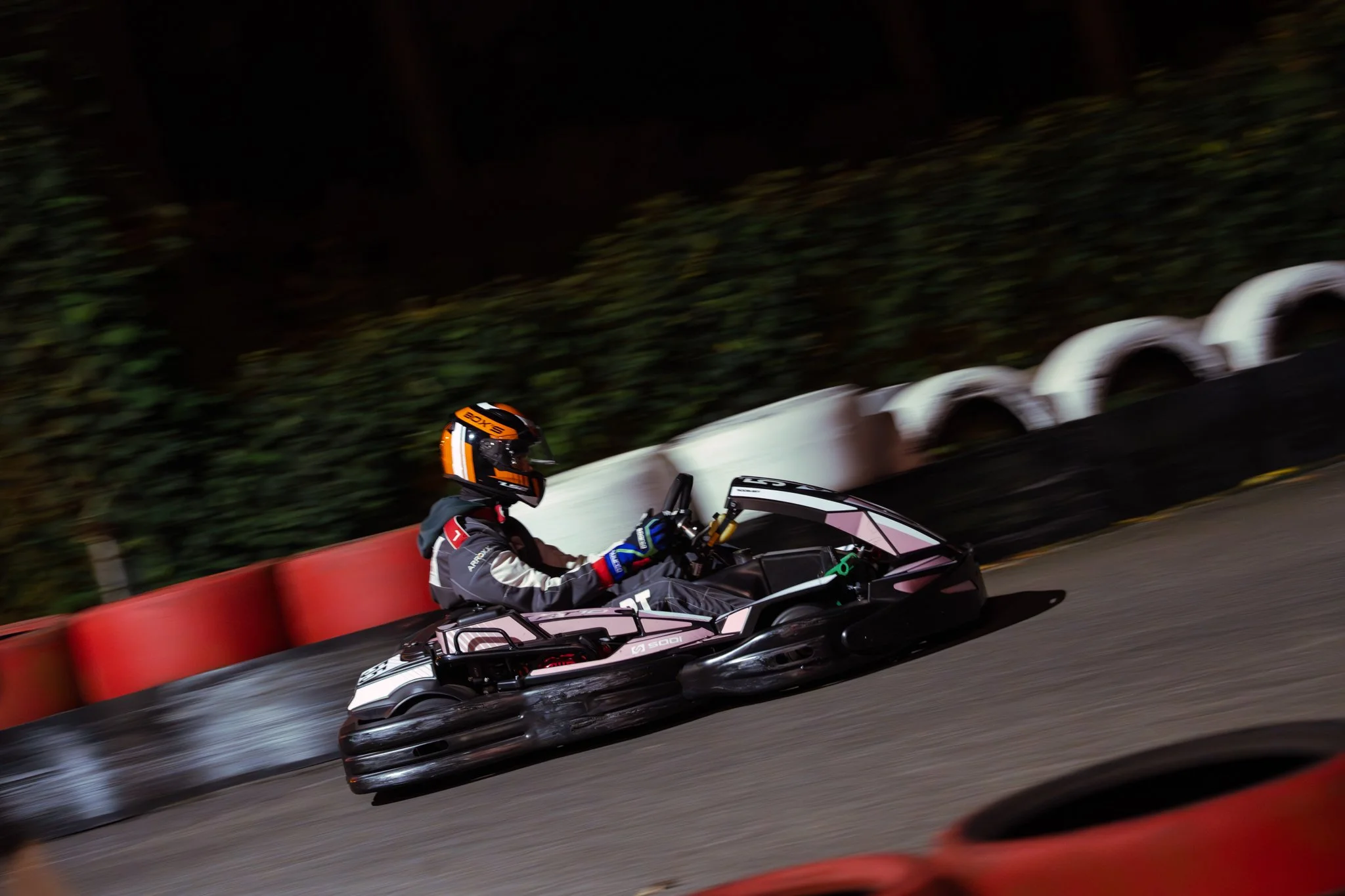 A person driving a go-kart on an outdoor track at night, wearing a helmet and racing gear, with red and black barriers along the track.