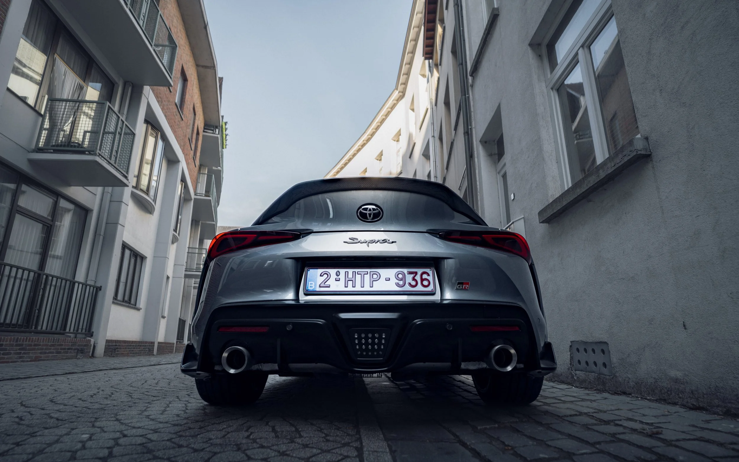 A rear view of a silver Toyota Supra parked between two buildings on a cobblestone street.