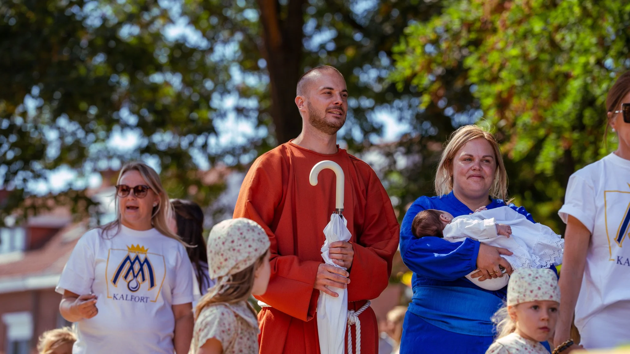 Group of people participating in an outdoor event with trees in the background. Among them is a man in a red robe holding a white umbrella, and a woman in a blue robe holding a baby. Other children and adults are present, some wearing casual clothes 
