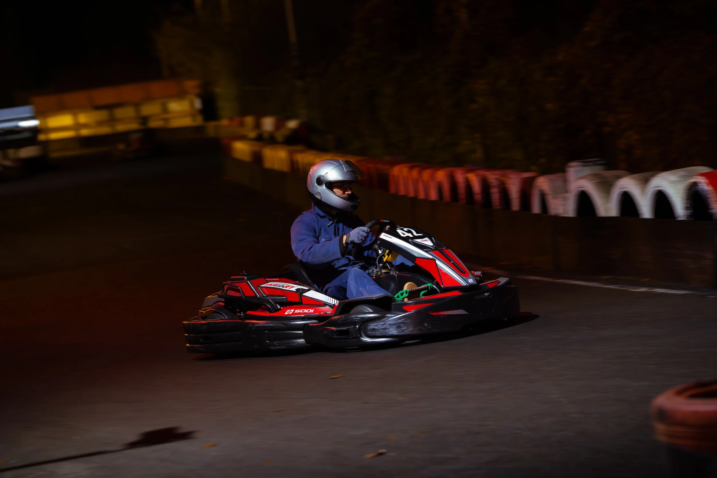 A person in a silver helmet and blue jacket riding a red and black go-kart at night, with a blurred background of a fence and trees.
