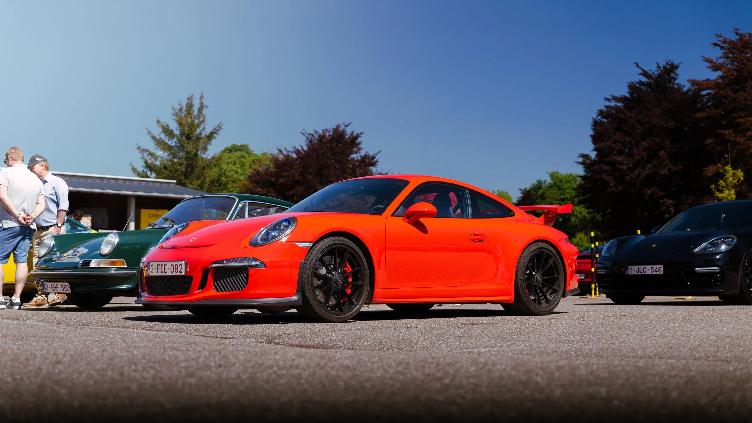 A red Porsche 911 sports car parked next to a black Porsche and vintage green car at a car show, with people in casual clothing and trees in the background on a sunny day.