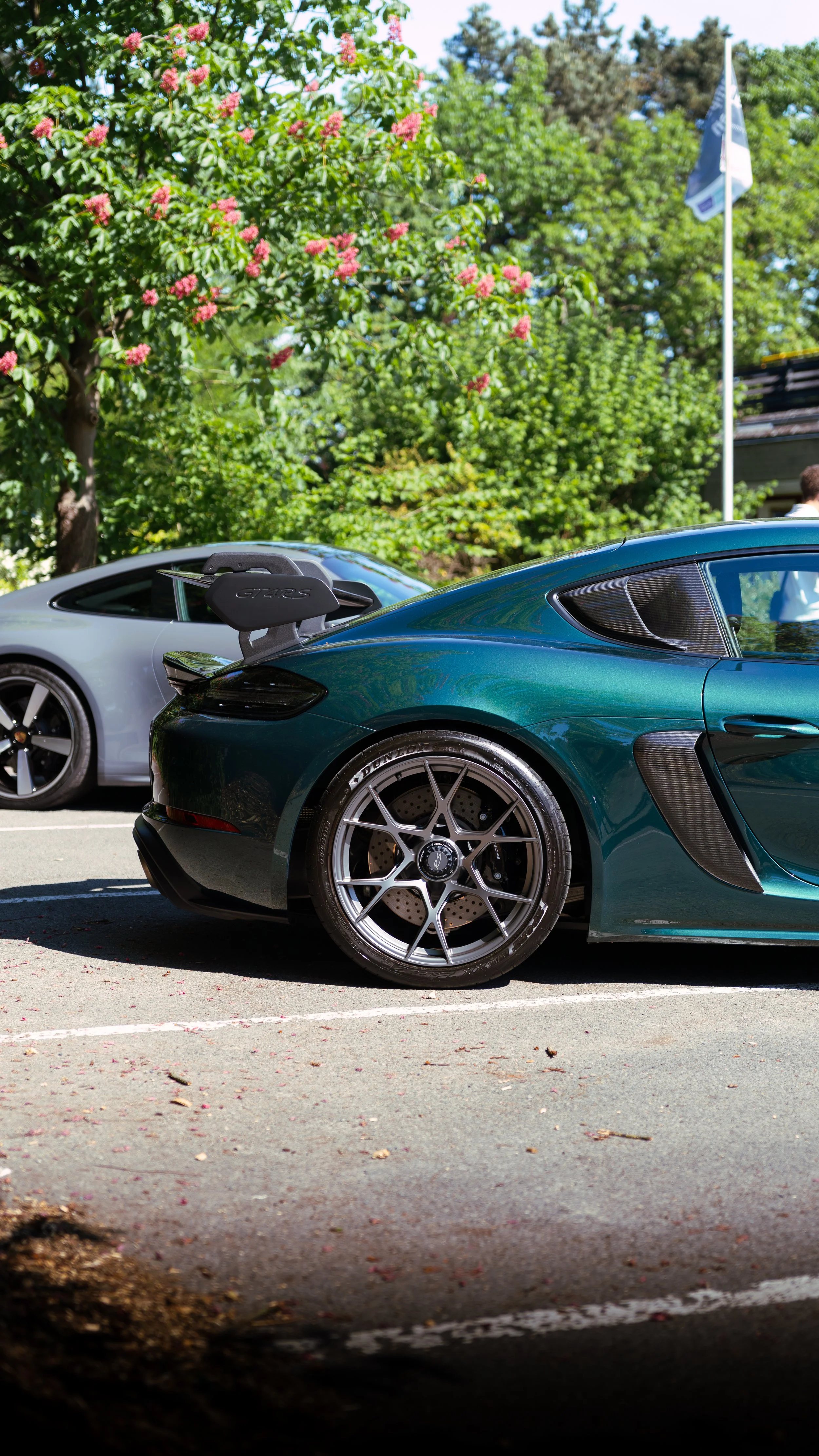 A green sports car parked next to a silver sports car in a parking lot surrounded by trees and greenery.