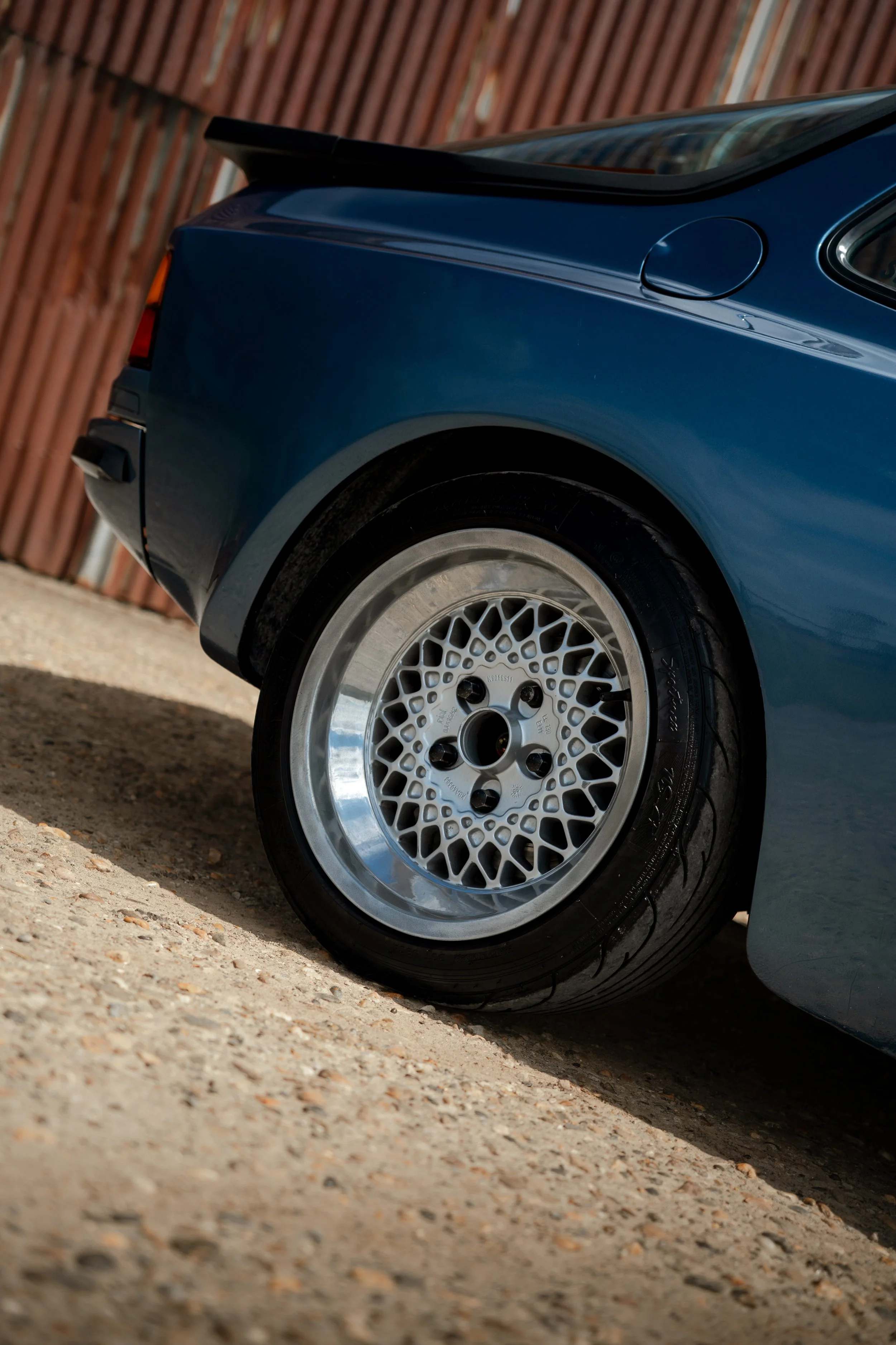 Close-up of the rear wheel and body of a blue car with a wire-spoke rim, parked on gravel with a wooden fence in the background.