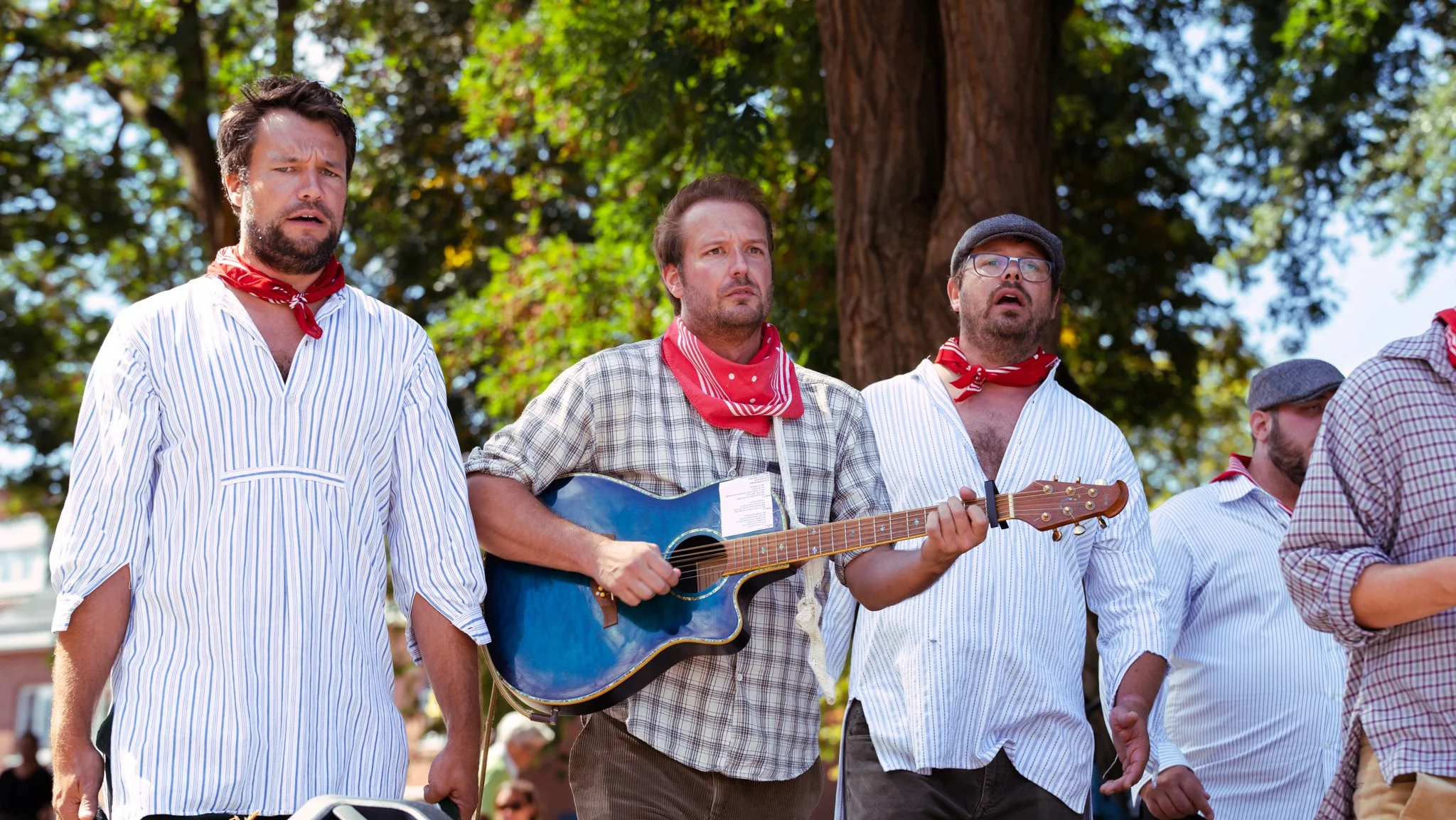 Group of men dressed in traditional Spanish attire standing outdoors, with one man playing guitar, under large trees in the background.