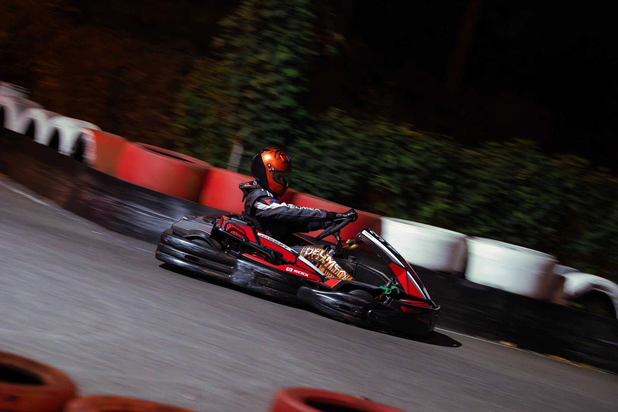 A person wearing a black and orange helmet and racing suit driving a go-kart on a track at night, with orange and white barriers along the side.
