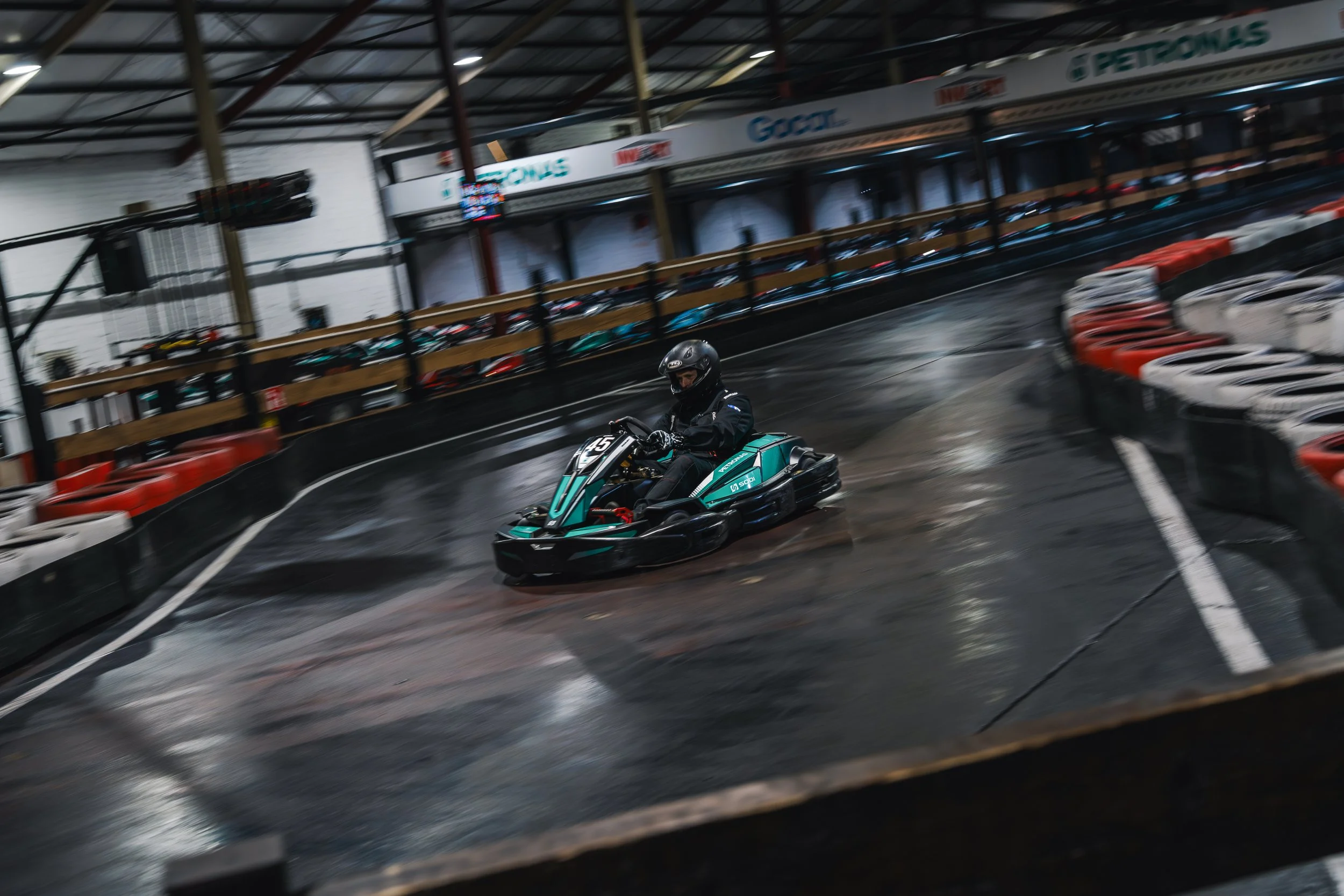 A person wearing a helmet driving a go-kart on an indoor race track with black and white barriers.