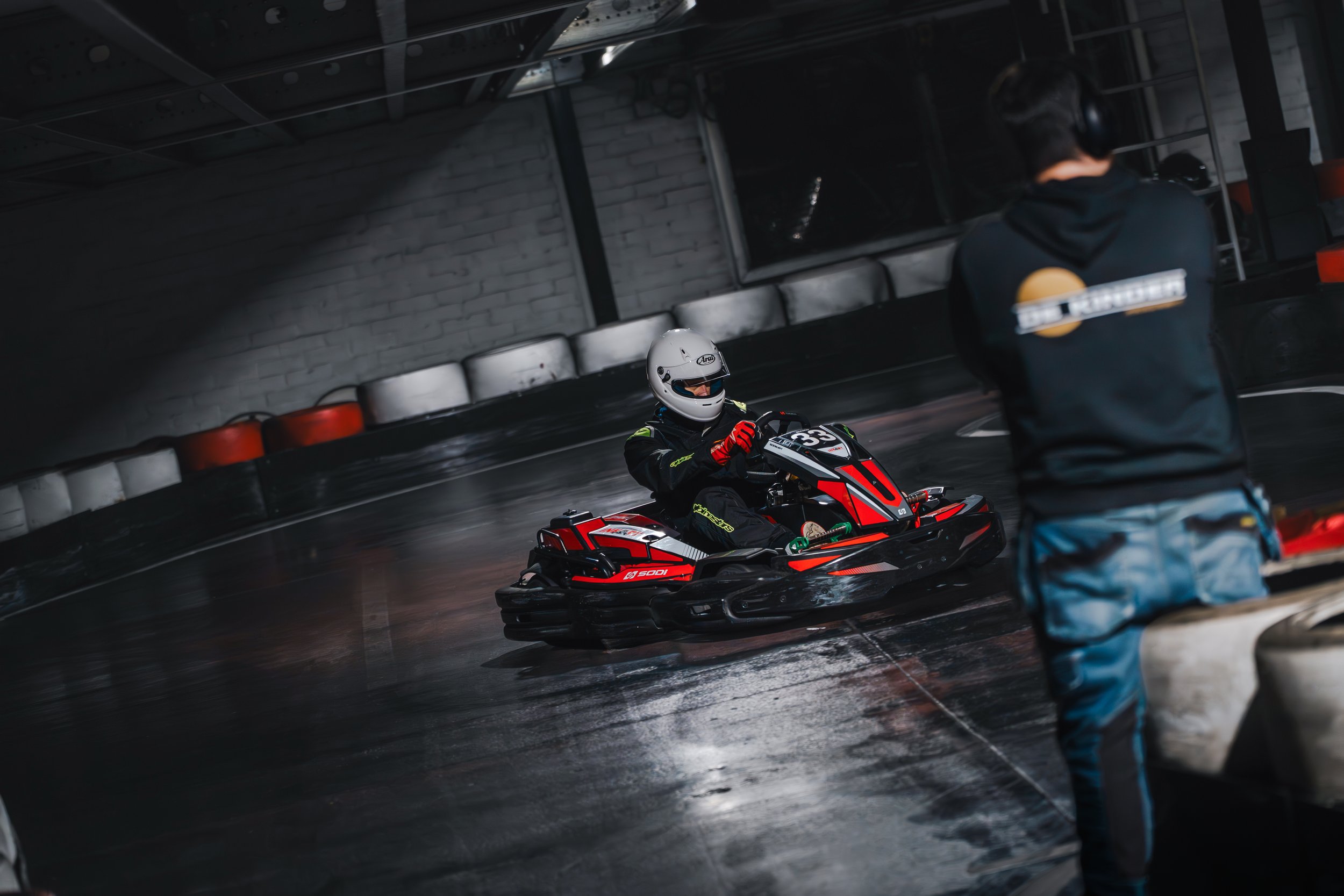 A person riding a go-kart wearing a white helmet, with another person standing nearby in a hoodie, inside an indoor go-kart racing facility with a brick wall and tires in the background.