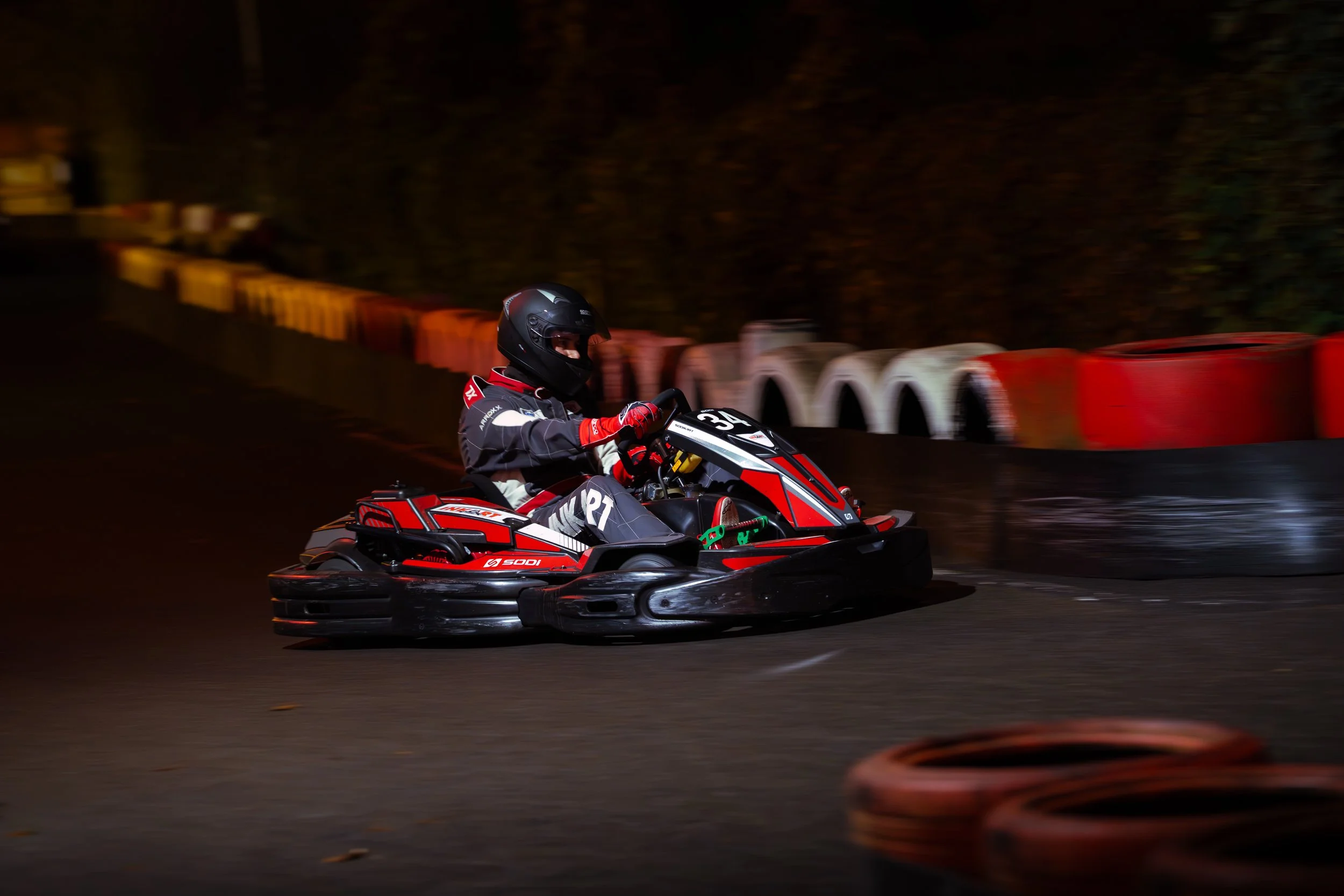 A person wearing a black helmet and racing suit riding a red and black go-kart on a dark race track at night, with orange and white barriers lining the track.