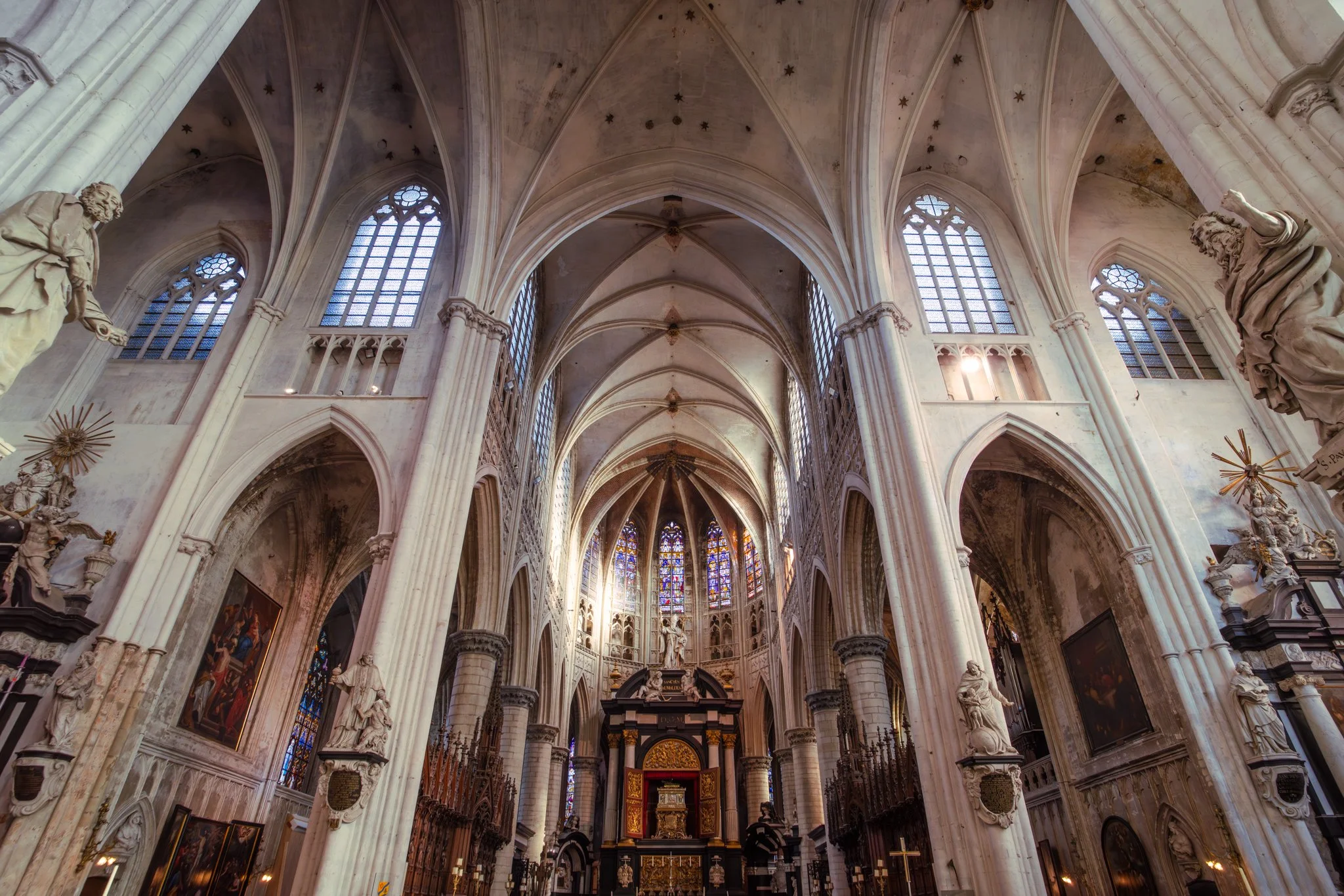Interior view of a Gothic cathedral showing tall arched ceilings, stained glass windows, and ornate statues along the walls.