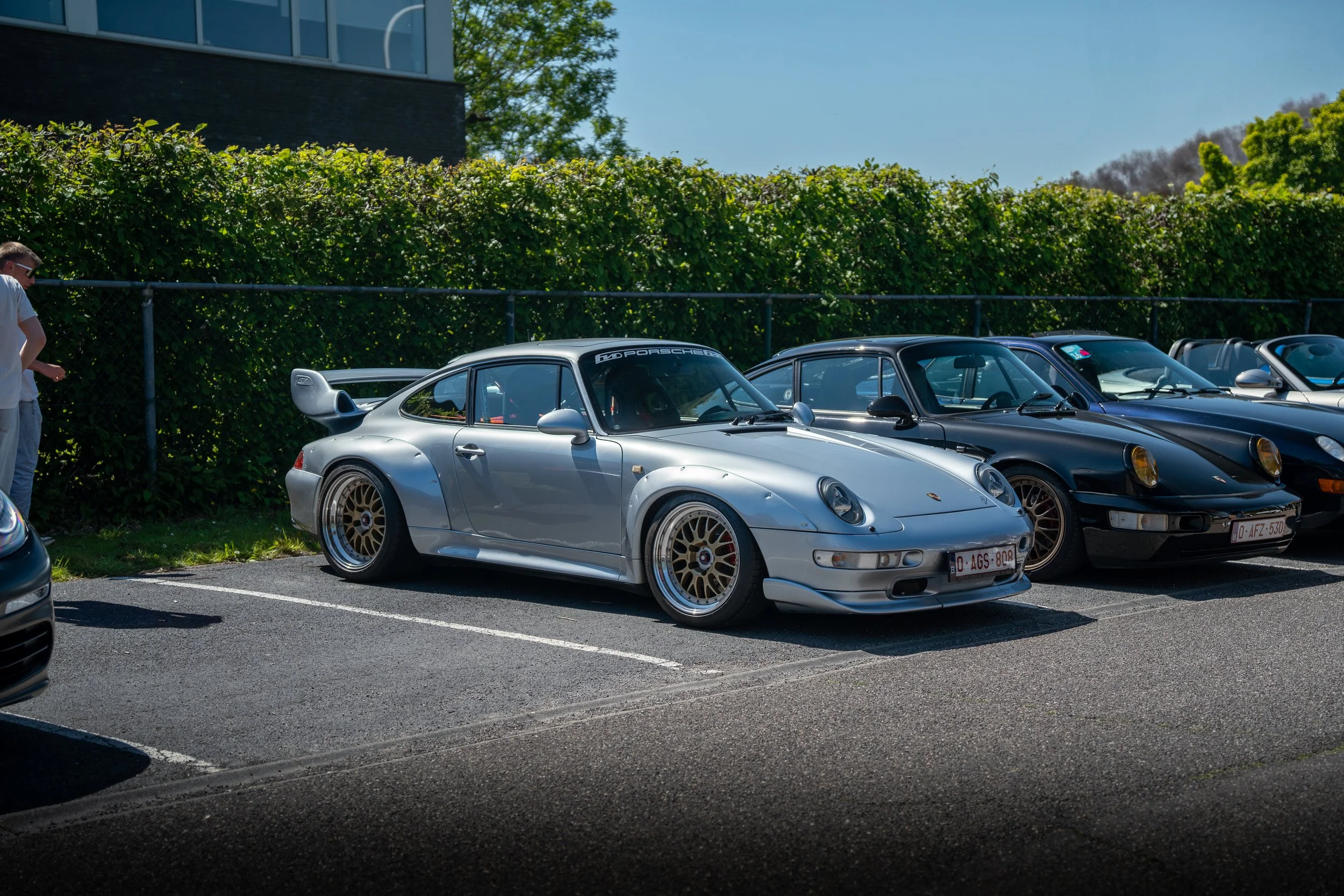 A silver Porsche 911 with gold wheels parked next to black Porsche 911s at a car show on a sunny day, with a fence and green trees in the background.