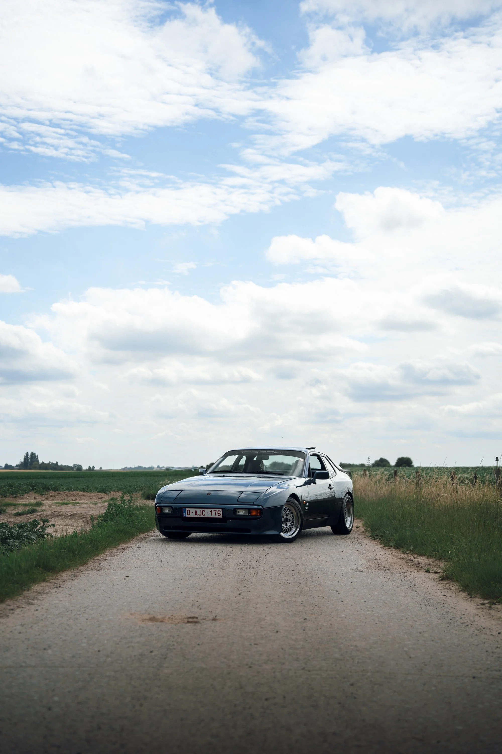 A black Porsche car parked on a small dirt road in a rural area, with fields and a partly cloudy sky in the background.