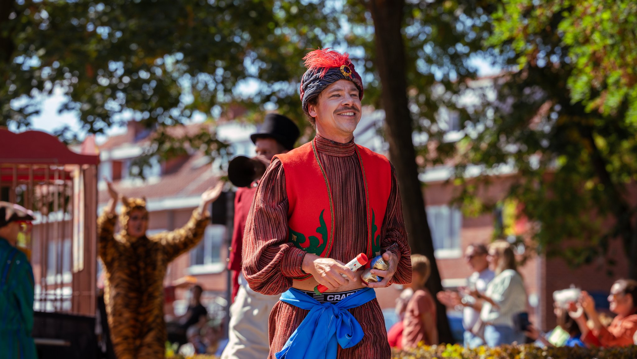 A man in colorful, traditional attire, with a red vest, patterned shirt, and a decorative headpiece with feathers, stands outdoors at a festival or parade with other people in costumes and casual clothing, and trees and buildings in the background.