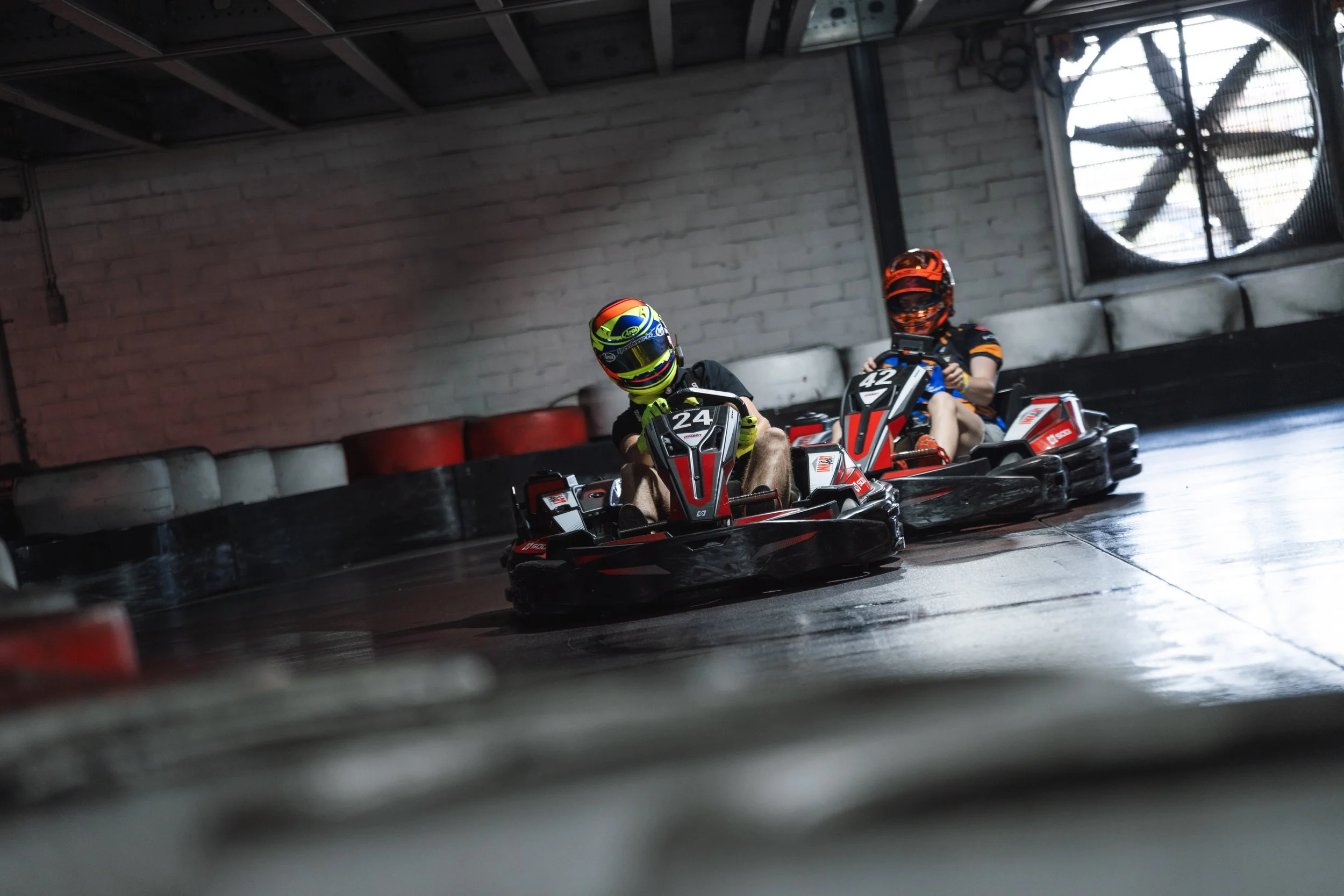 Two people wearing helmets are riding go-karts indoors near a brick wall and a large industrial fan.