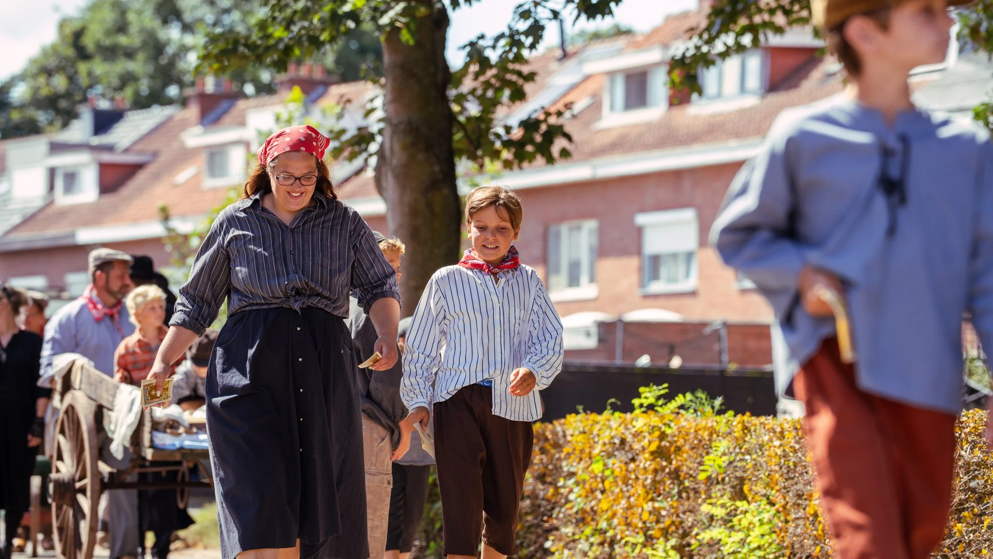 People participating in a traditional outdoor festival, dressed in historical costumes, walking along a street lined with trees and houses, holding cards or booklets.