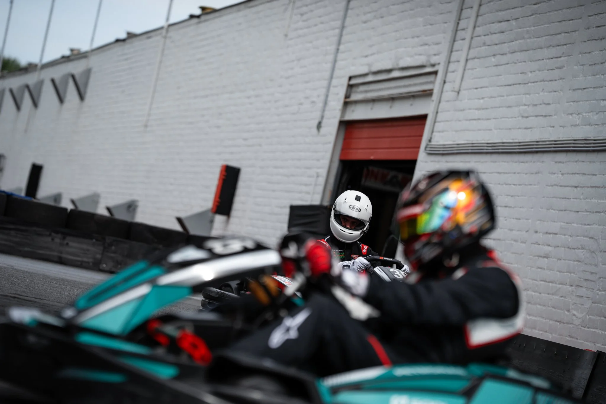 Two motorcyclists in racing gear and helmets on a race track, with one rider looking at the other inside a garage or obstacle area with a white brick wall in the background.