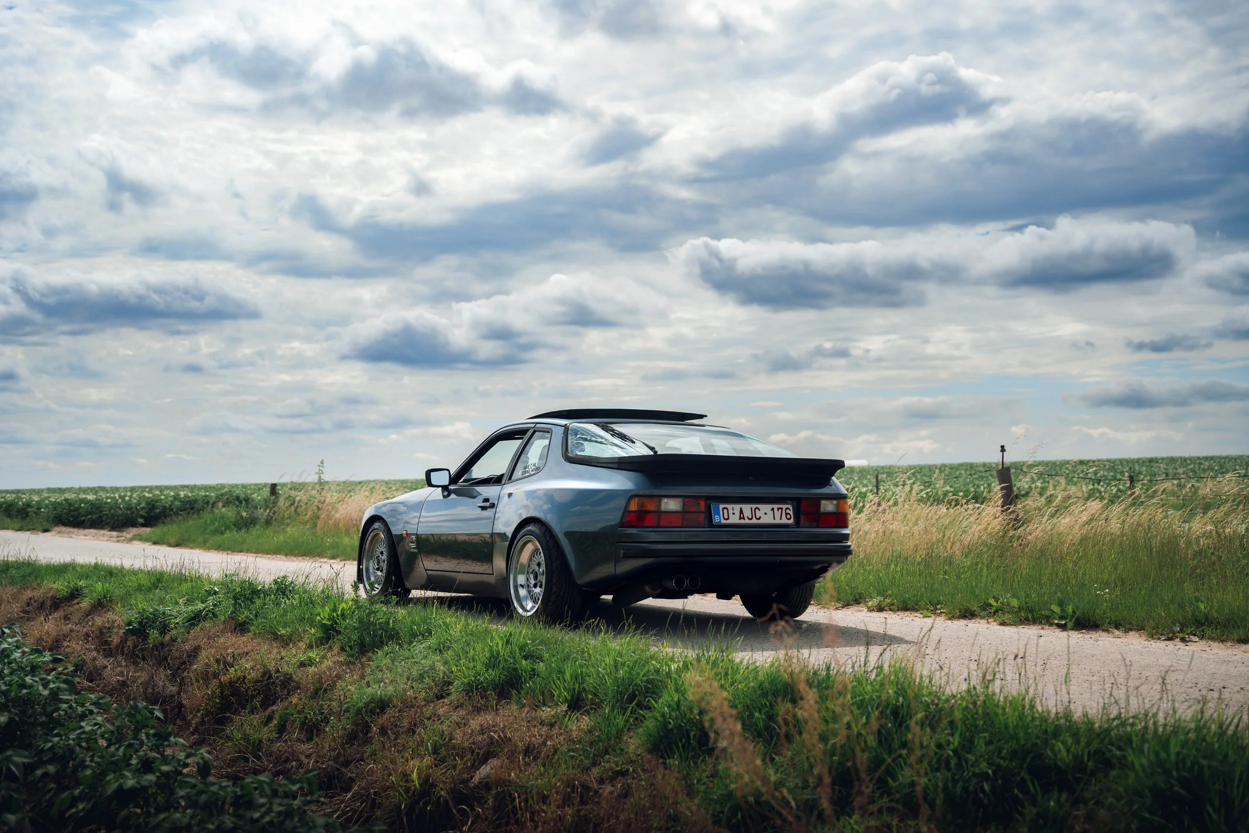 A vintage silver sports car parked by the side of a country road, with green fields and a cloudy sky in the background.