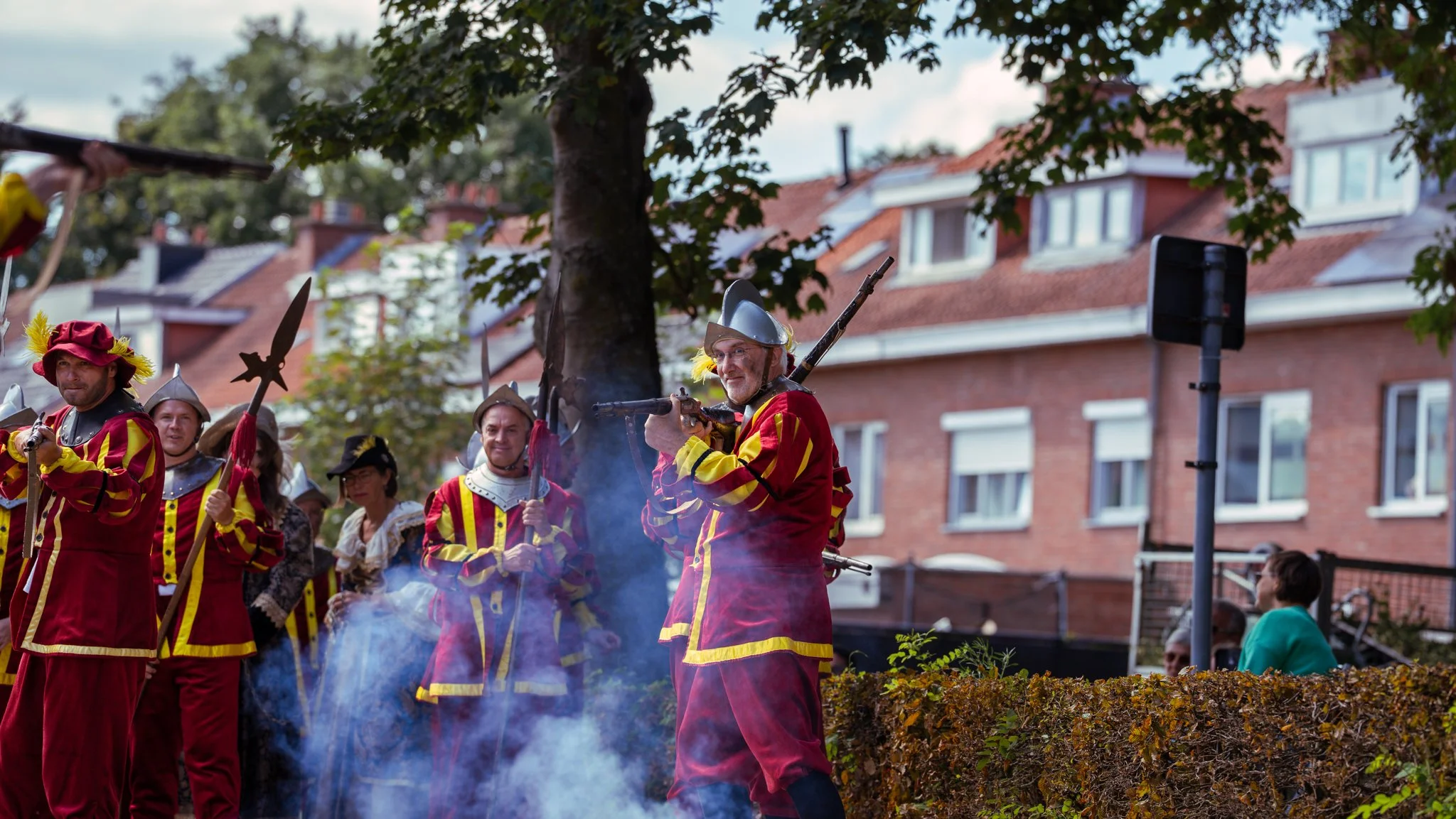 People dressed in colorful, historical costumes participating in a parade or festival on a city street, with some holding spears or weapons, surrounded by a crowd and trees, with red brick buildings in the background.
