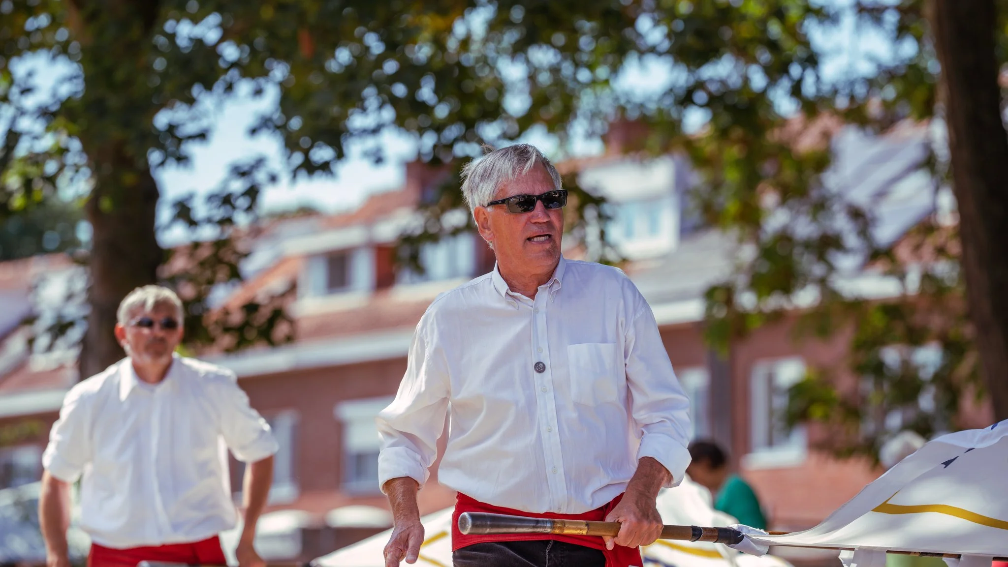 Older man in white shirt and sunglasses holding a flagpole at a parade or outdoor event, with another man in similar attire in the background, trees, and houses visible.