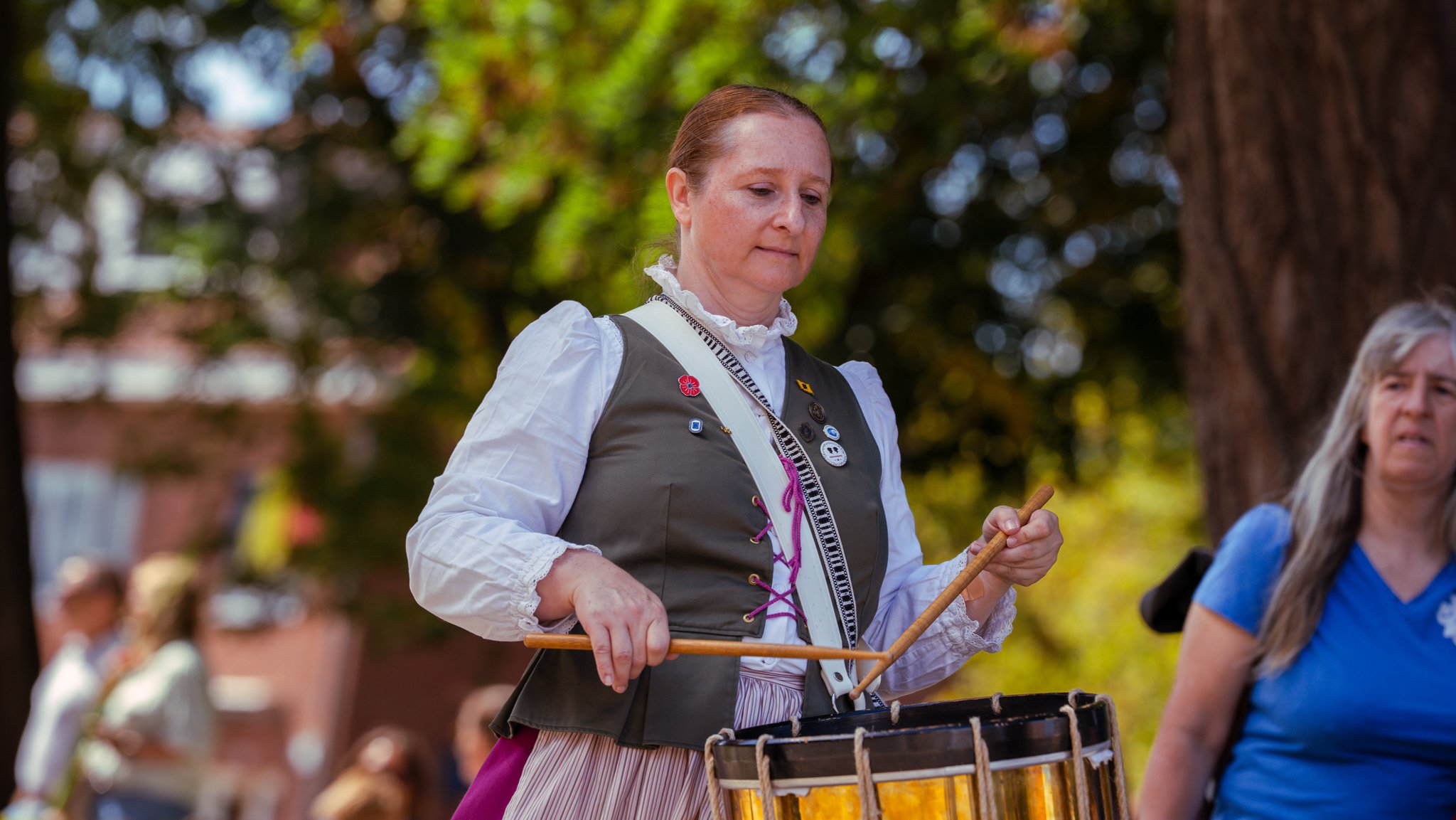 A woman with red hair playing a percussion instrument outdoors, wearing a vintage-style outfit with buttons and pins, while other people are in the background.
