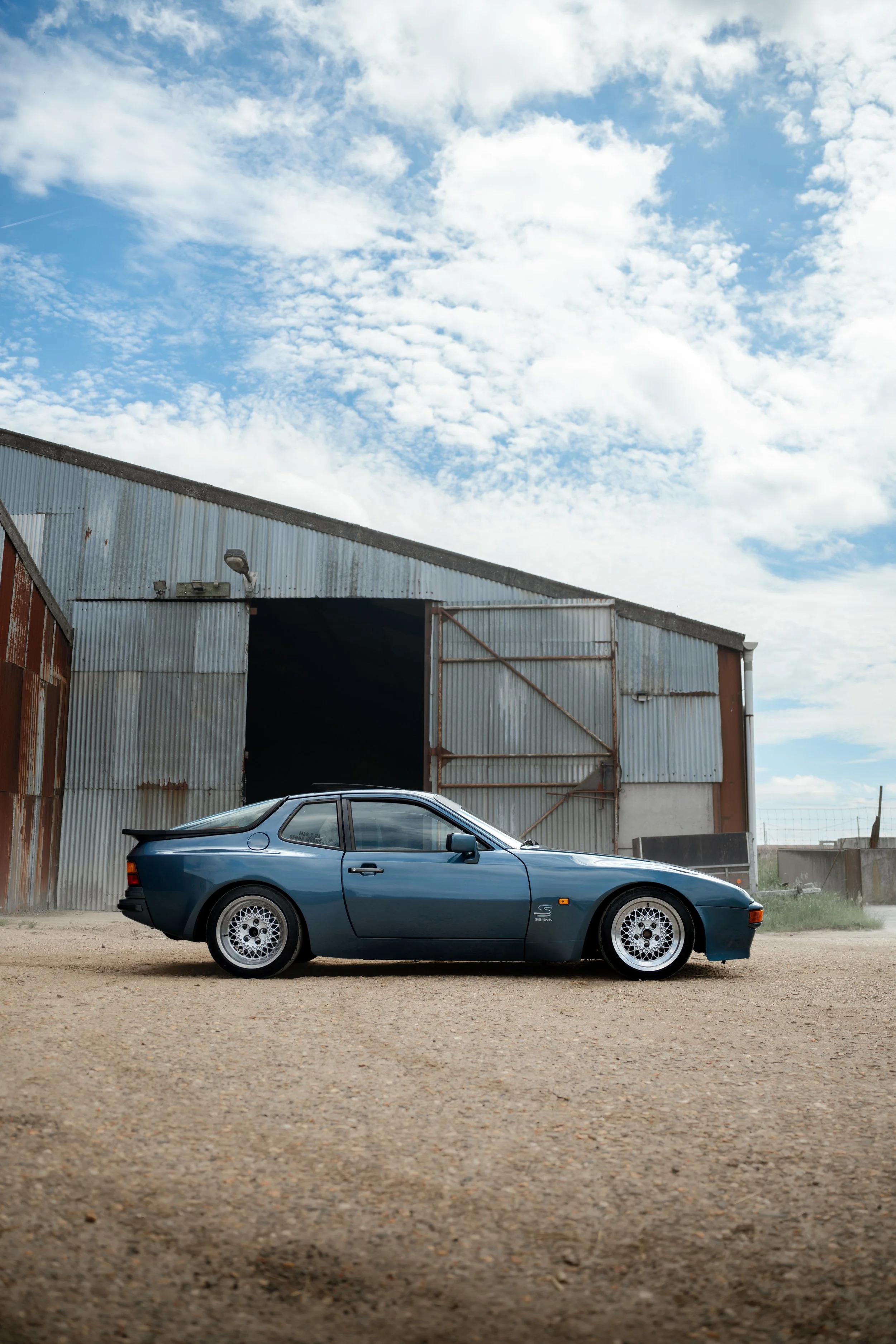 A blue vintage sports car with pop-up headlights parked on a dirt ground in front of a rustic metal building with an open door and a streetlamp on a cloudy day.