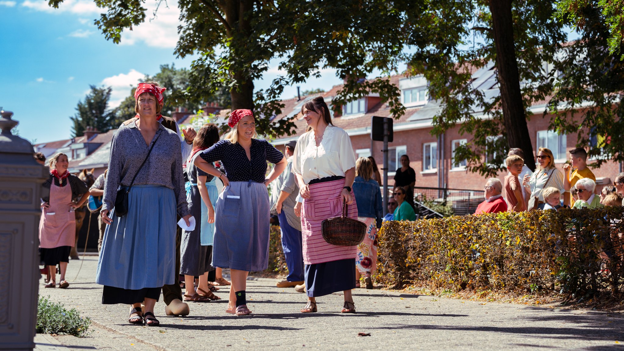 People dressed in traditional clothing gathered outdoors, engaging in conversation, with some sitting on benches and others standing under trees in a sunny neighborhood.