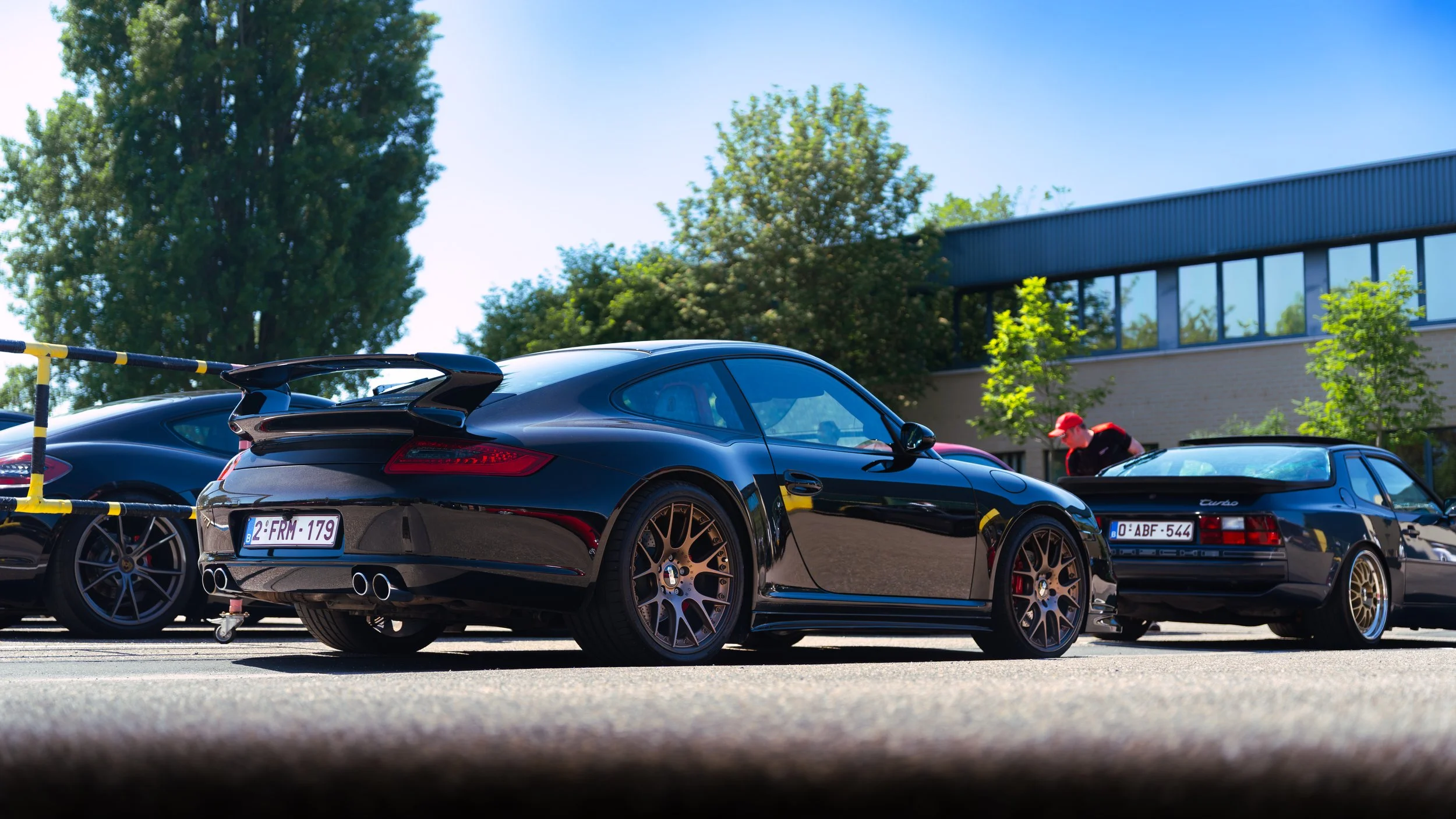 Black Porsche 911 with a rear spoiler parked in a lot next to two other classic cars, with a person in a red hat inspecting the car. There are trees and a building in the background.