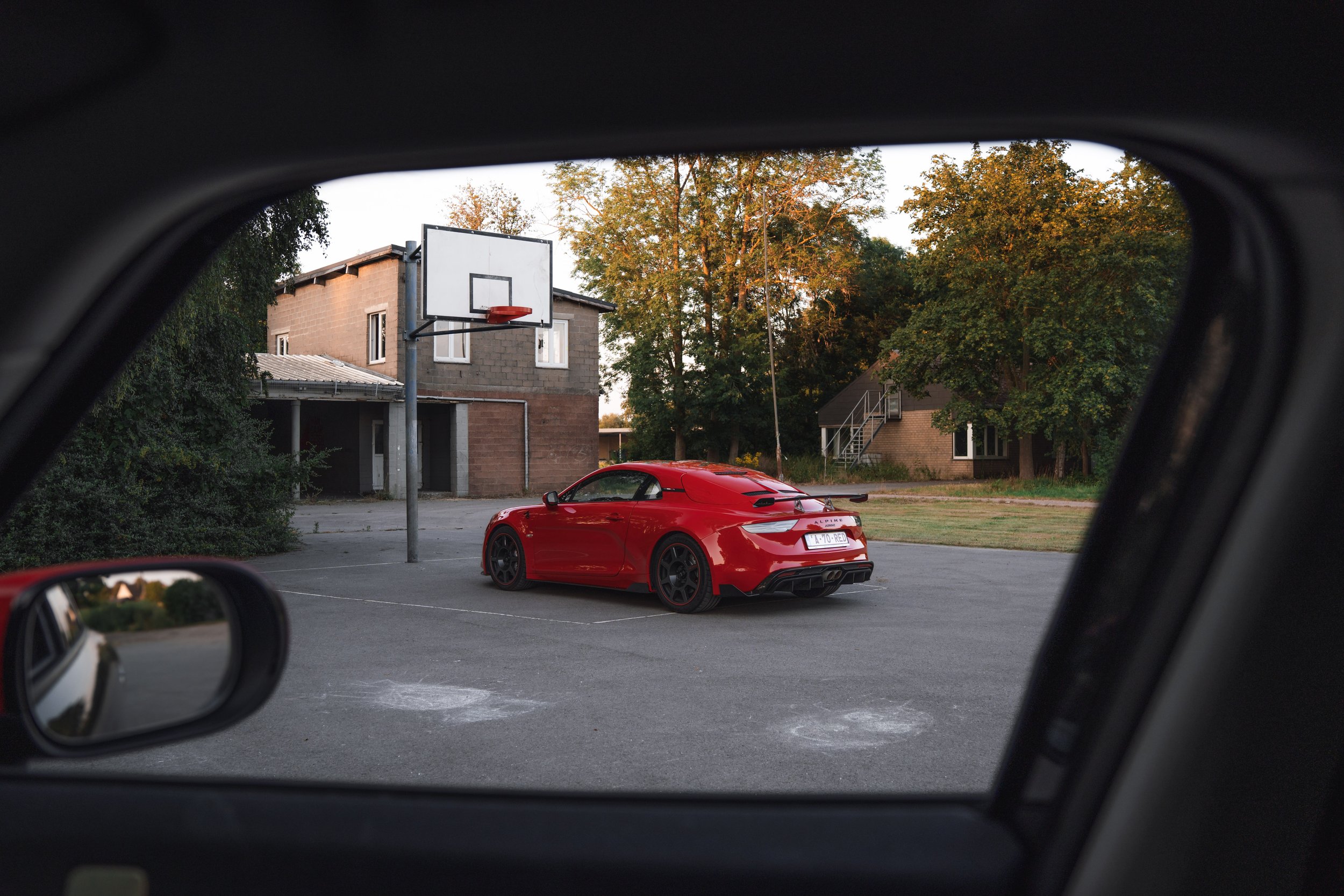 Red sports car parked near a basketball hoop outside houses with trees in the background, viewed from inside a vehicle.