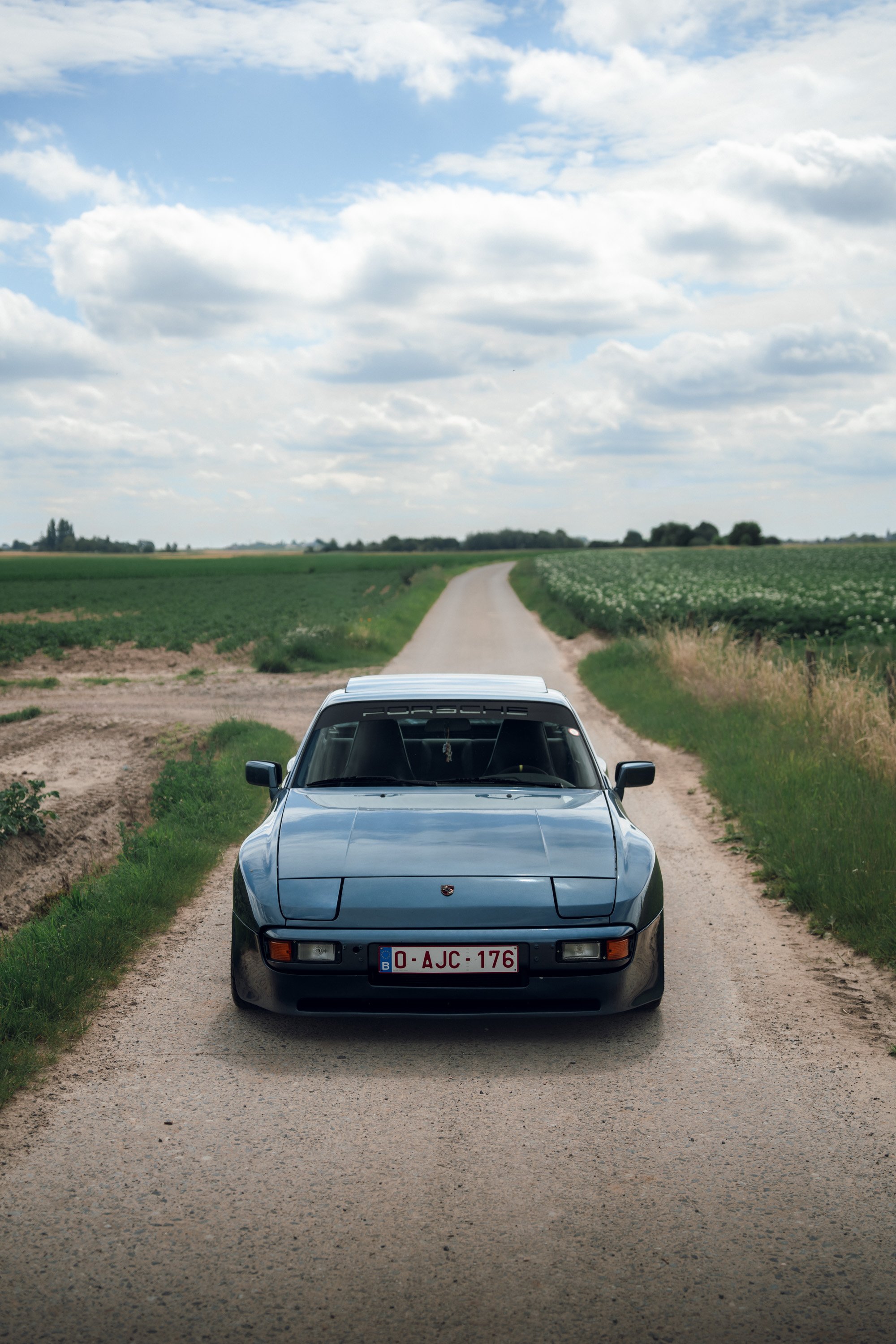 A silver Porsche sports car parked on a rural dirt road between fields of green crops, with a sky filled with white clouds overhead.
